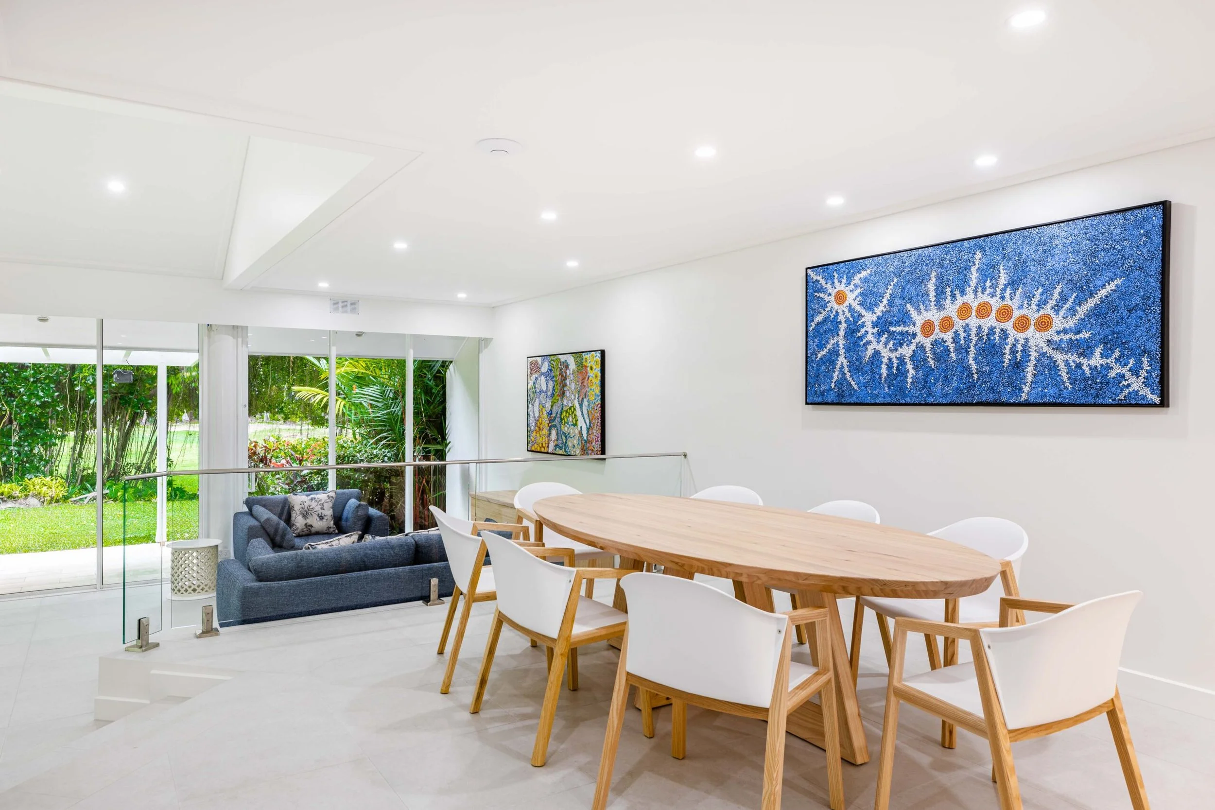 Modern living room with white walls, recessed lighting, a wooden dining table with white and wooden chairs, a blue sofa, and large glass sliding doors leading to a garden with greenery.