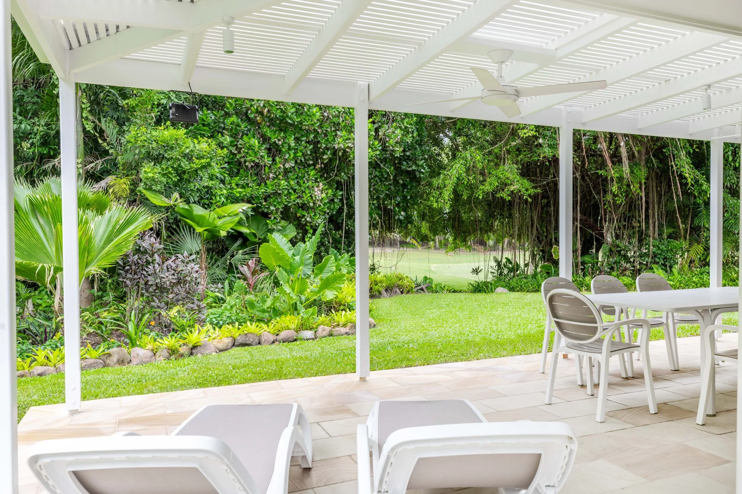 White outdoor patio with tables and chairs, covered by a white pergola, overlooking a lush green garden with various plants and trees.