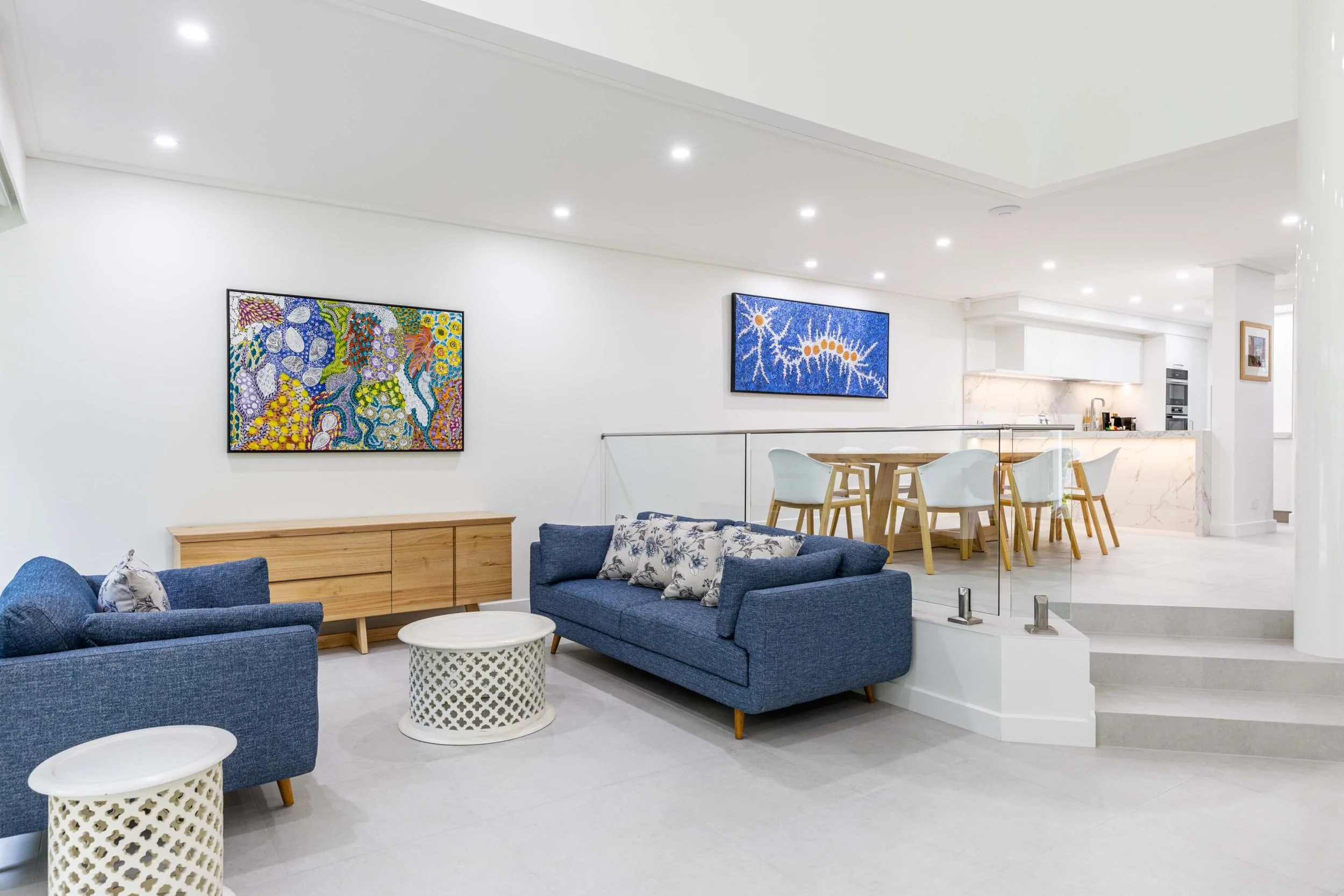 A modern living room with two blue sofas, white and patterned throw pillows, and white side tables. Artwork hangs on white walls, and there is a dining area with a wooden table and white chairs, adjacent to a kitchen with white cabinetry and stainles