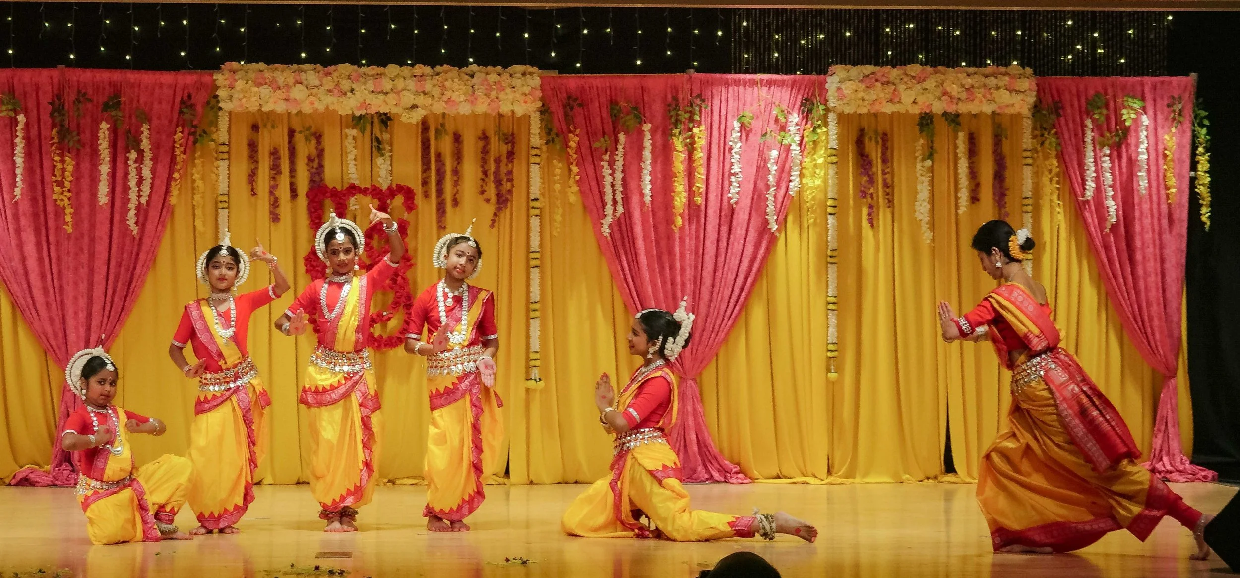 Children performing a traditional Indian dance on stage, dressed in colorful costumes with jewelry, in front of a decorated backdrop of yellow and pink curtains with floral decorations.