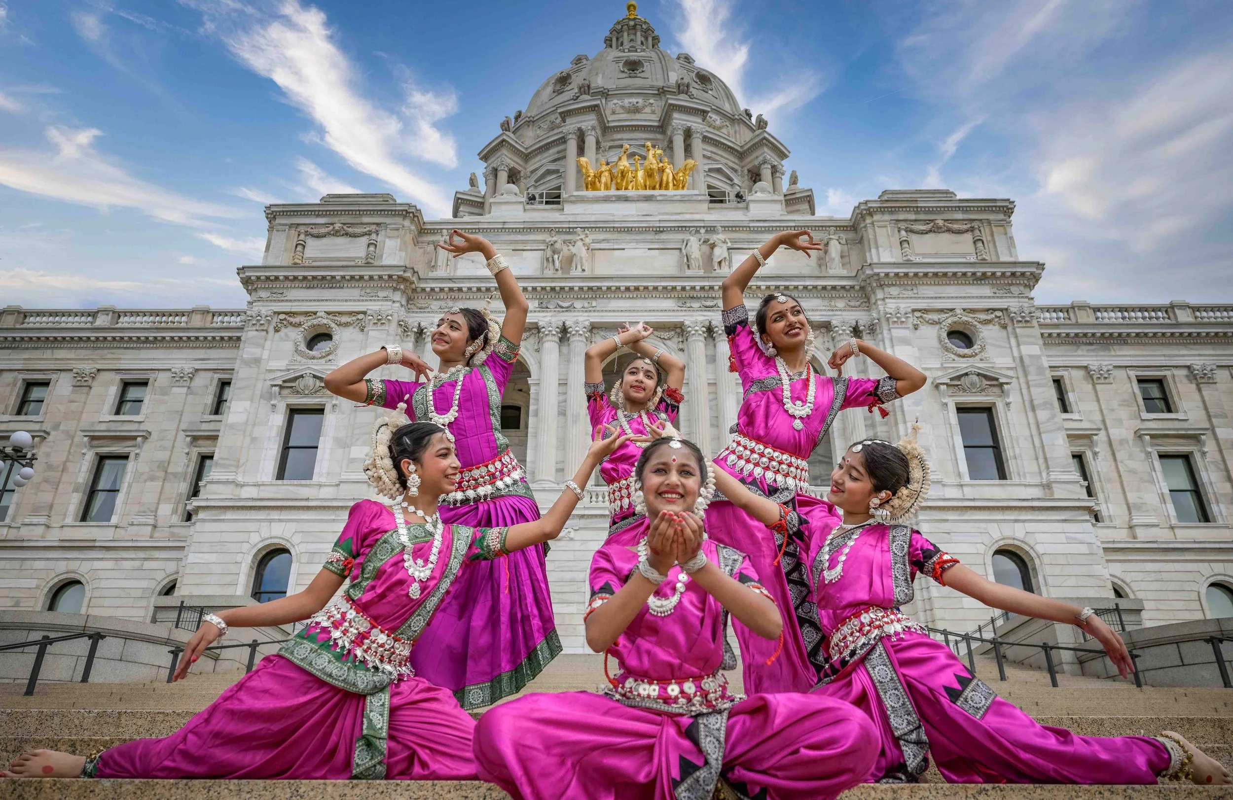 Group of young women in traditional pink Indian dance costumes performing a dance in front of a large white historic building with a dome and gold sculptures.