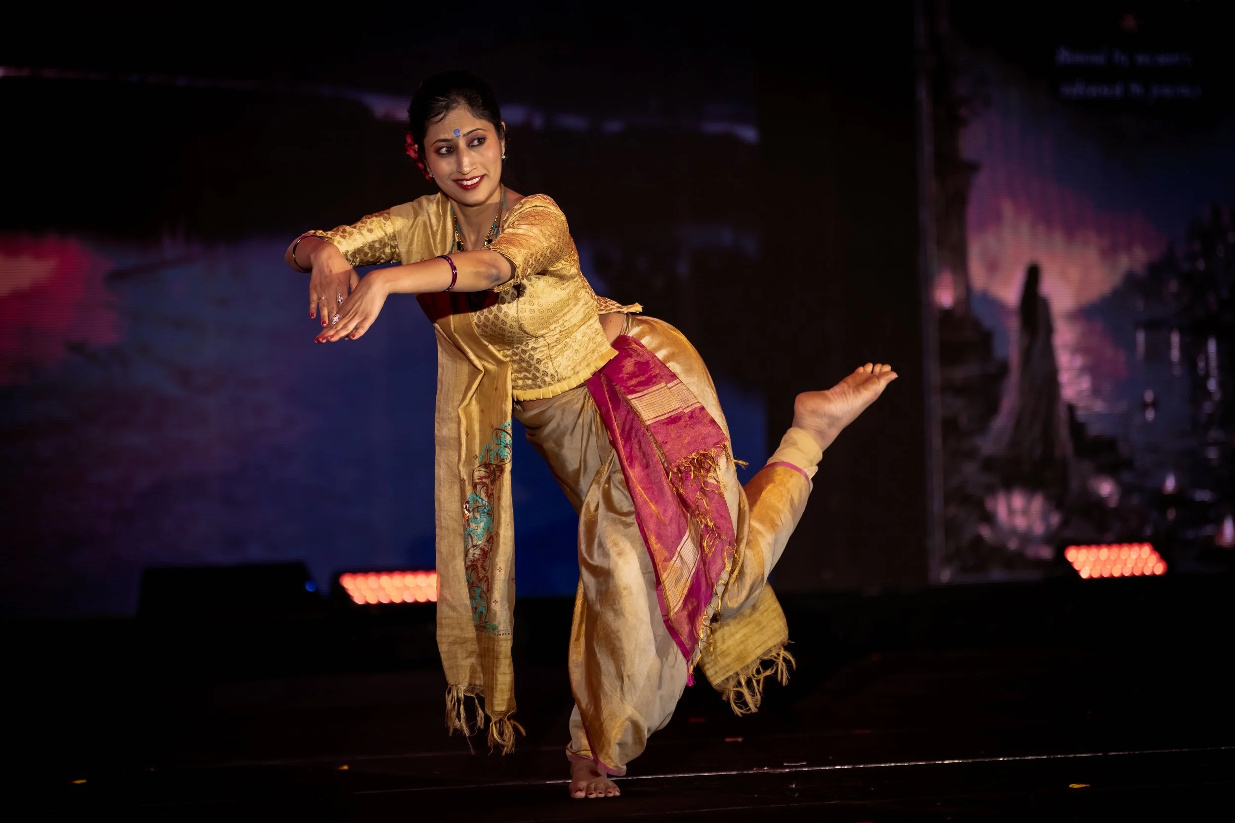 A woman performing a classical dance on stage, dressed in traditional Indian attire with a gold and pink sari, with her hair styled and makeup applied, captured during a graceful dance pose.