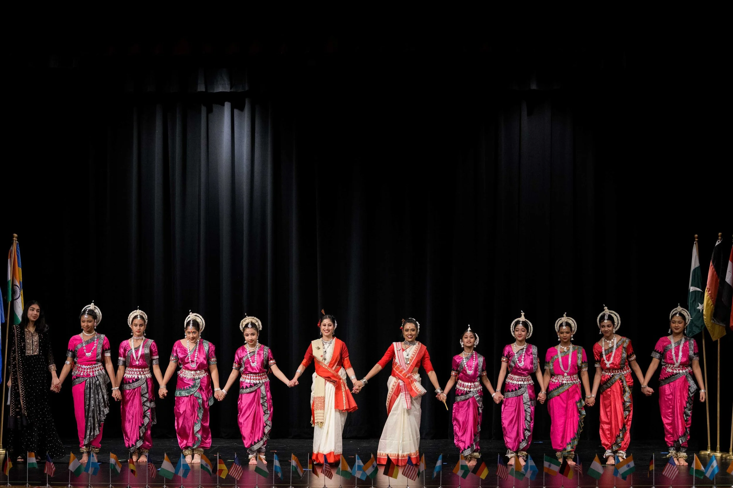 Group of women dressed in traditional Indian attire, holding hands on stage during a cultural dance performance.