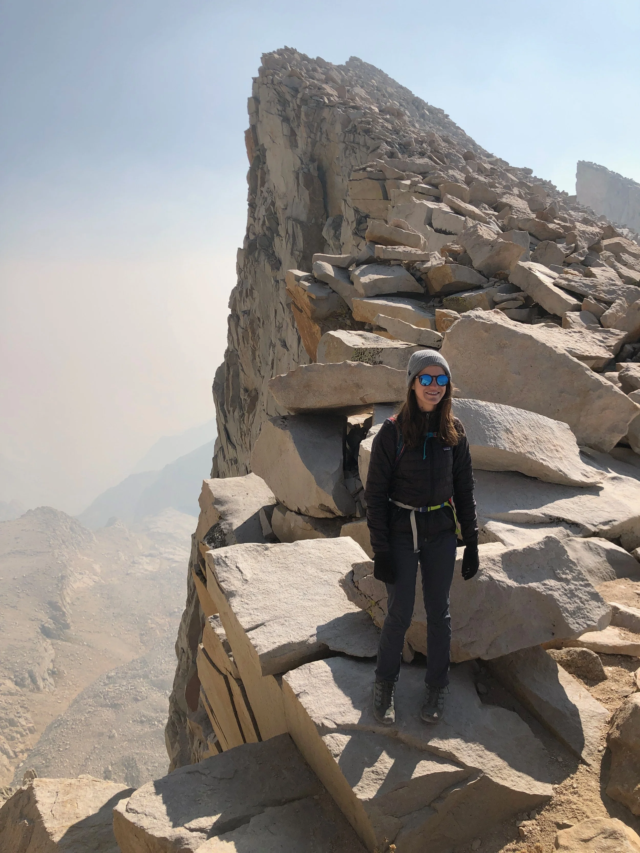 A woman standing on a rocky mountain ledge, surrounded by large boulders and a steep cliff, wearing outdoor gear including a beanie, sunglasses, and a jacket.
