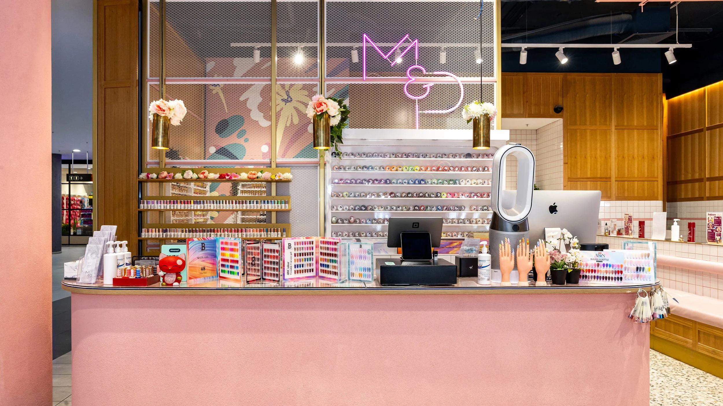 Beauty nail salon reception area with pink counter, display of nail polish bottles, color samples, a Dyson fan, desktop computer, and decorative flowers, with pink, gold, and wood accents.