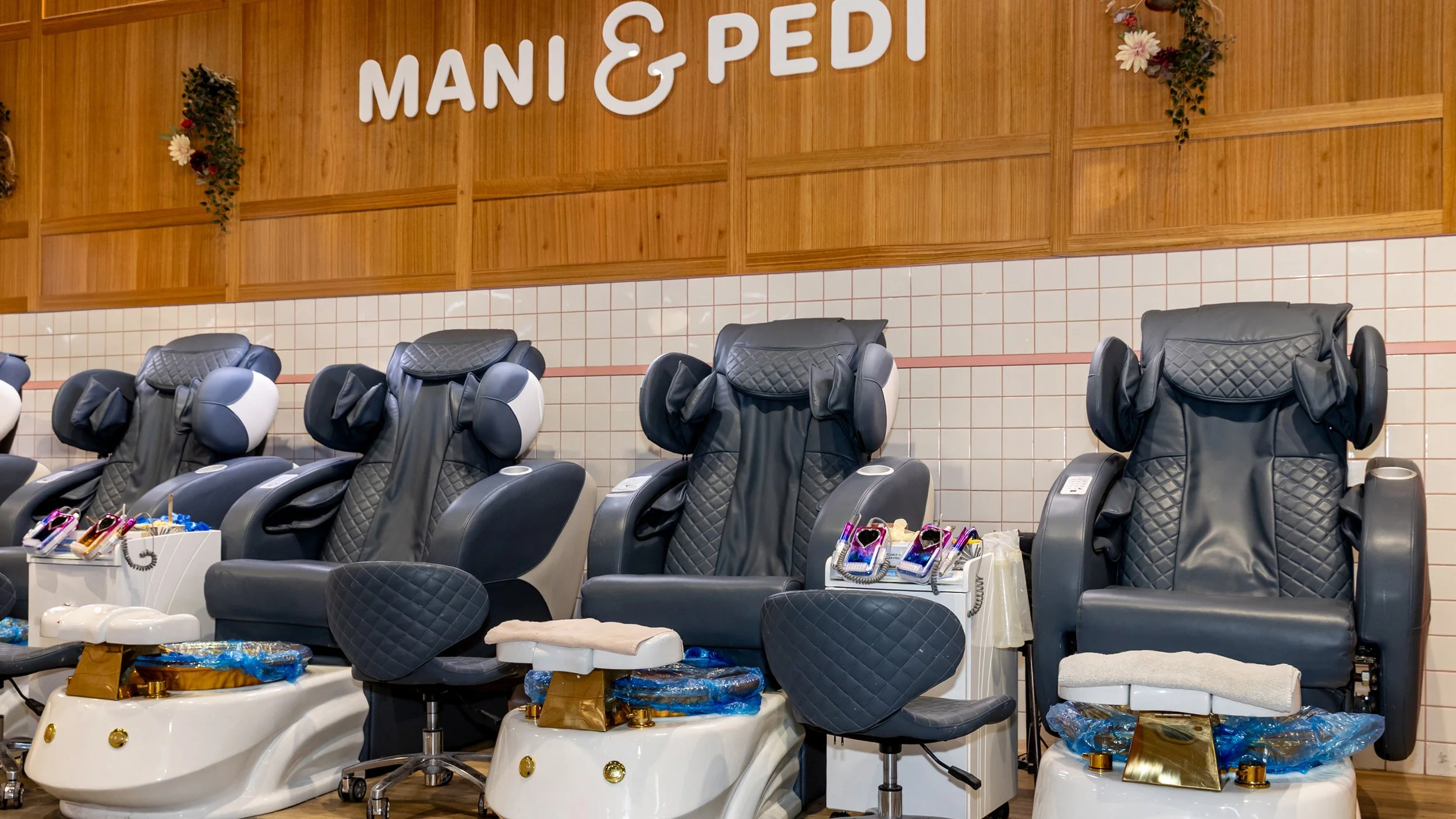 Row of massage chairs in a spa or salon, with arm and leg massagers and control buttons, located in front of a wooden wall with the sign 'Mani & Pedi' and decorative flowers.