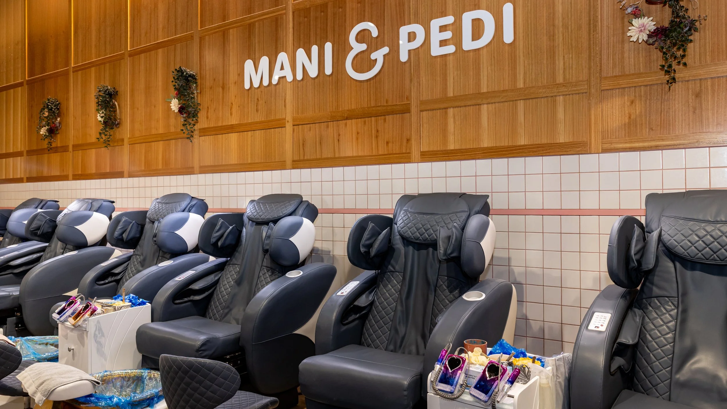 Spa area with massage chairs arranged in a row, each with a control panel and headrest. The wall above features a wooden panel with the sign 'MANI & PEDI' in white letters, and decorative flowers are mounted on the wooden wall.