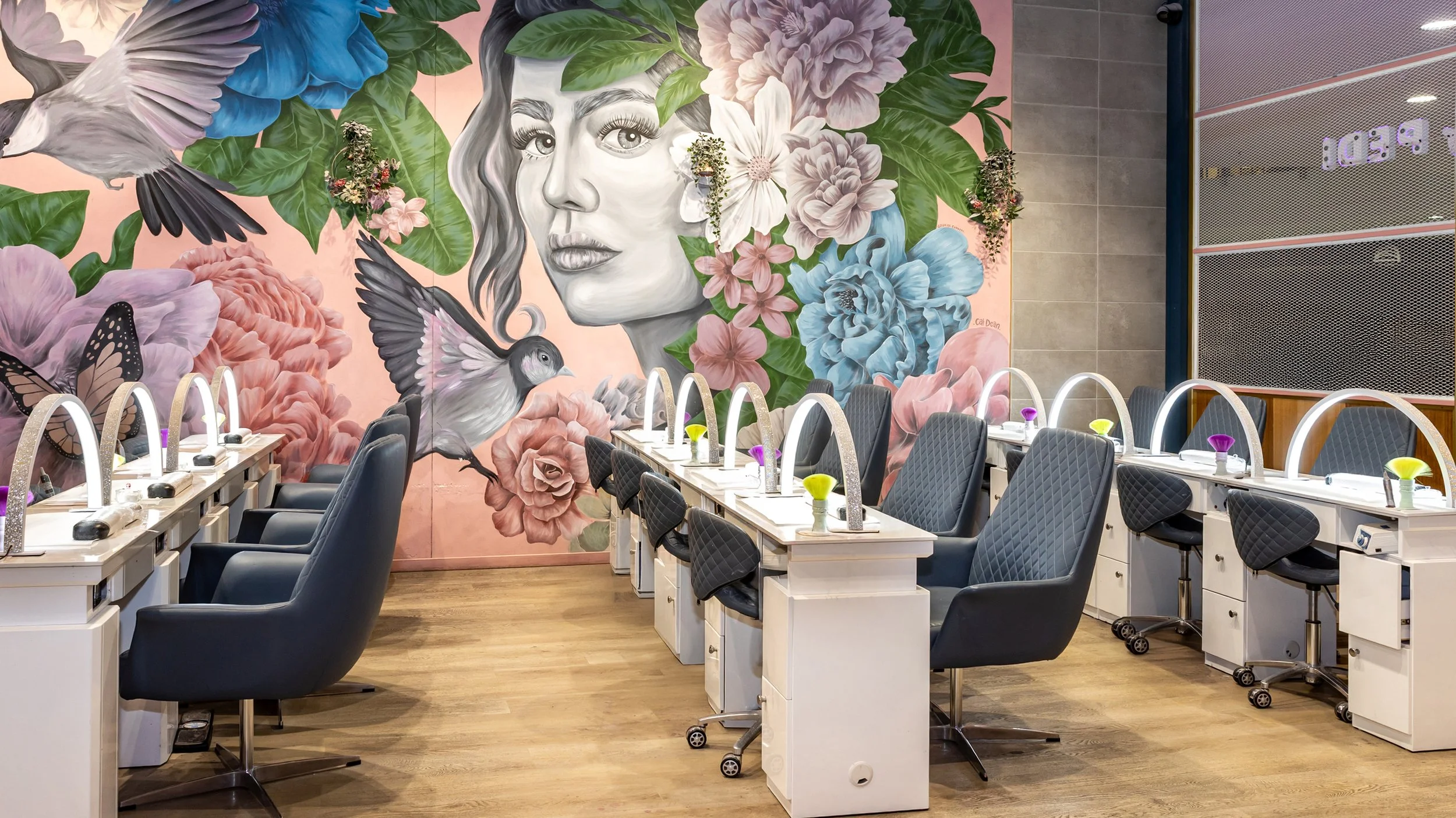 Beauty salon with nail stations, black chairs, and a large mural of a woman's face surrounded by flowers and birds on the wall.