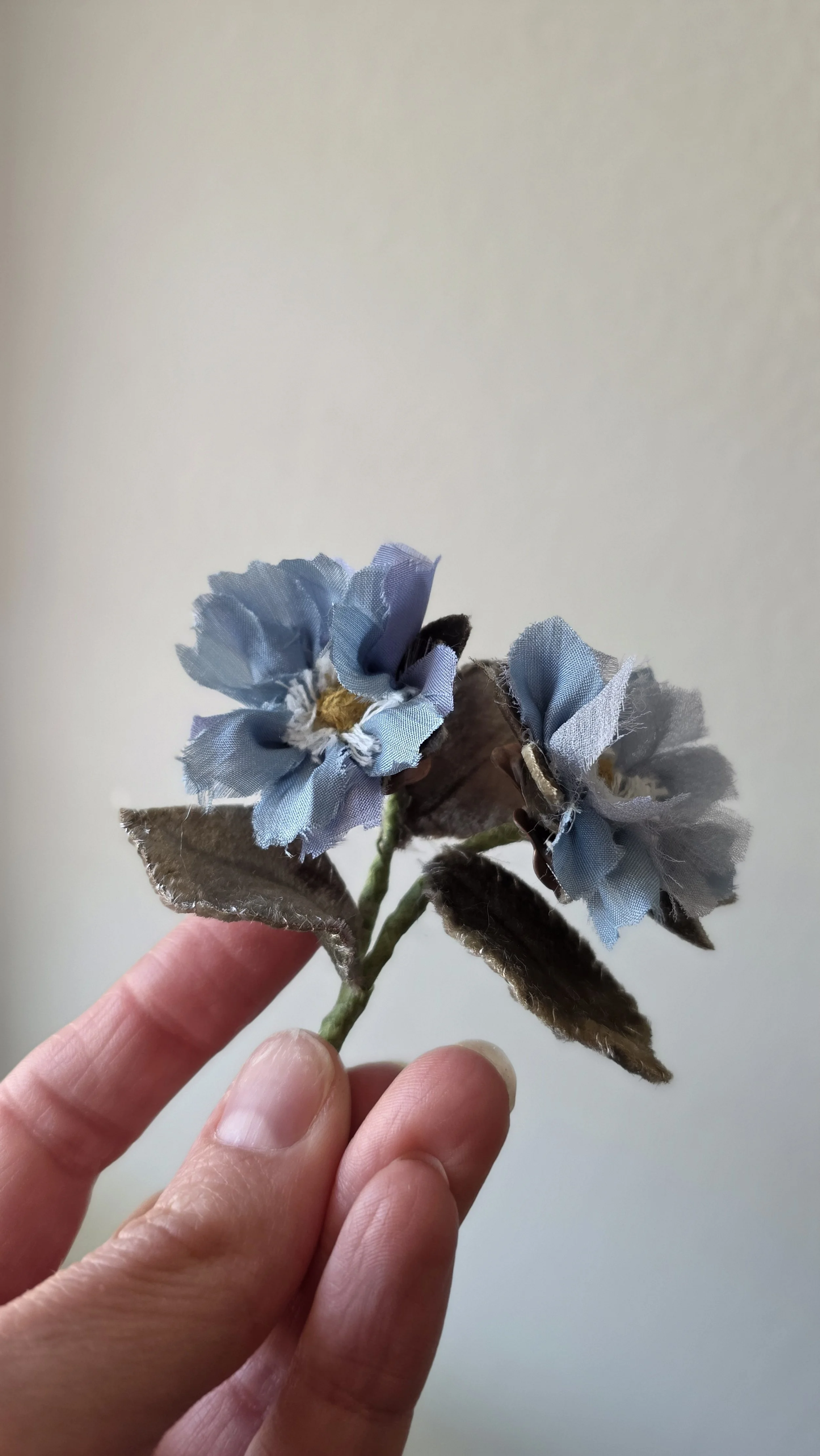 A hand holding a silk and velvet flower brooch with two blue silk flowers and green velvet leaves against a plain background.