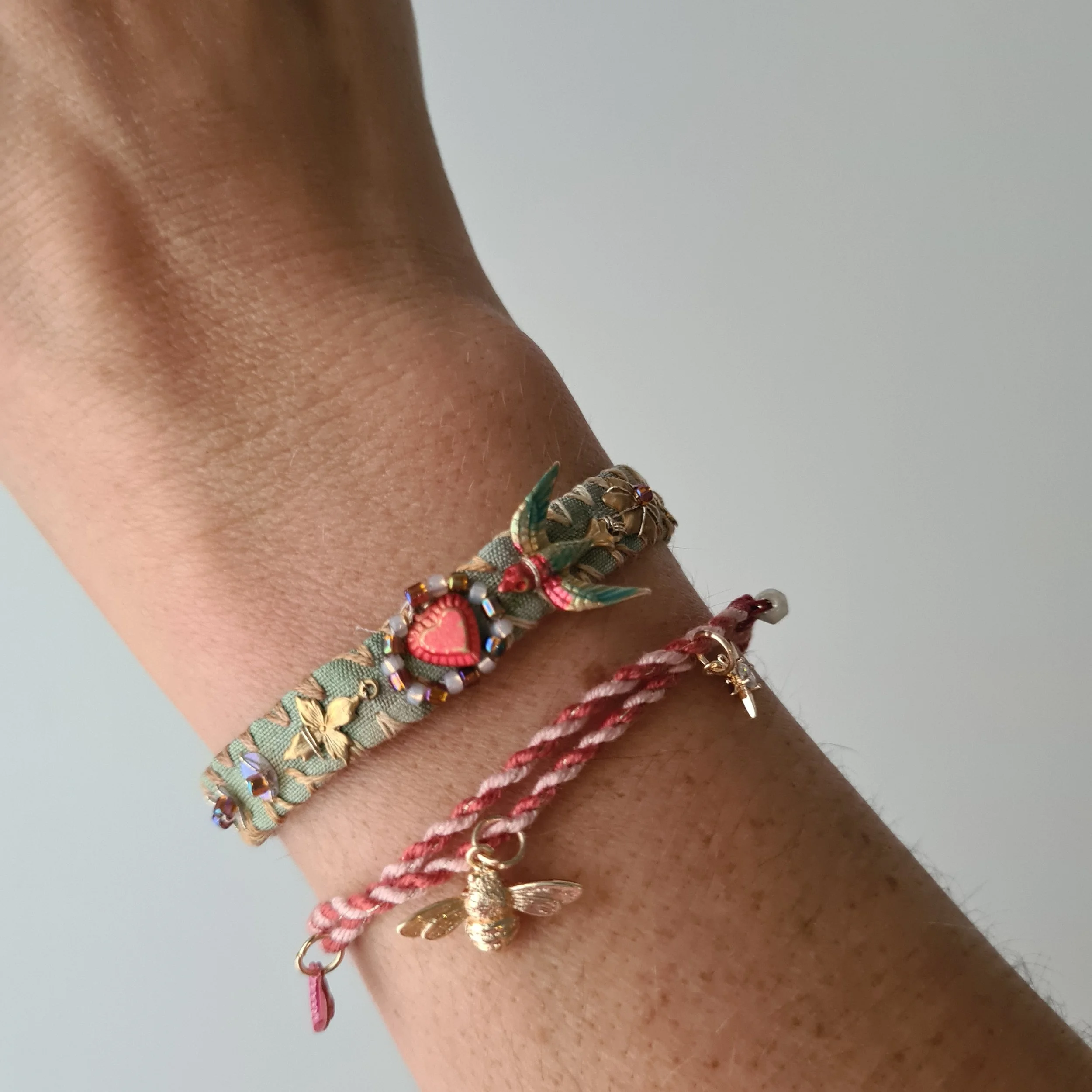 A person's wrist wearing two bracelets, one is an embroidered silk bracelet with a floral and bead design and the other is a pink and white twisted cord with a gold bee charm.