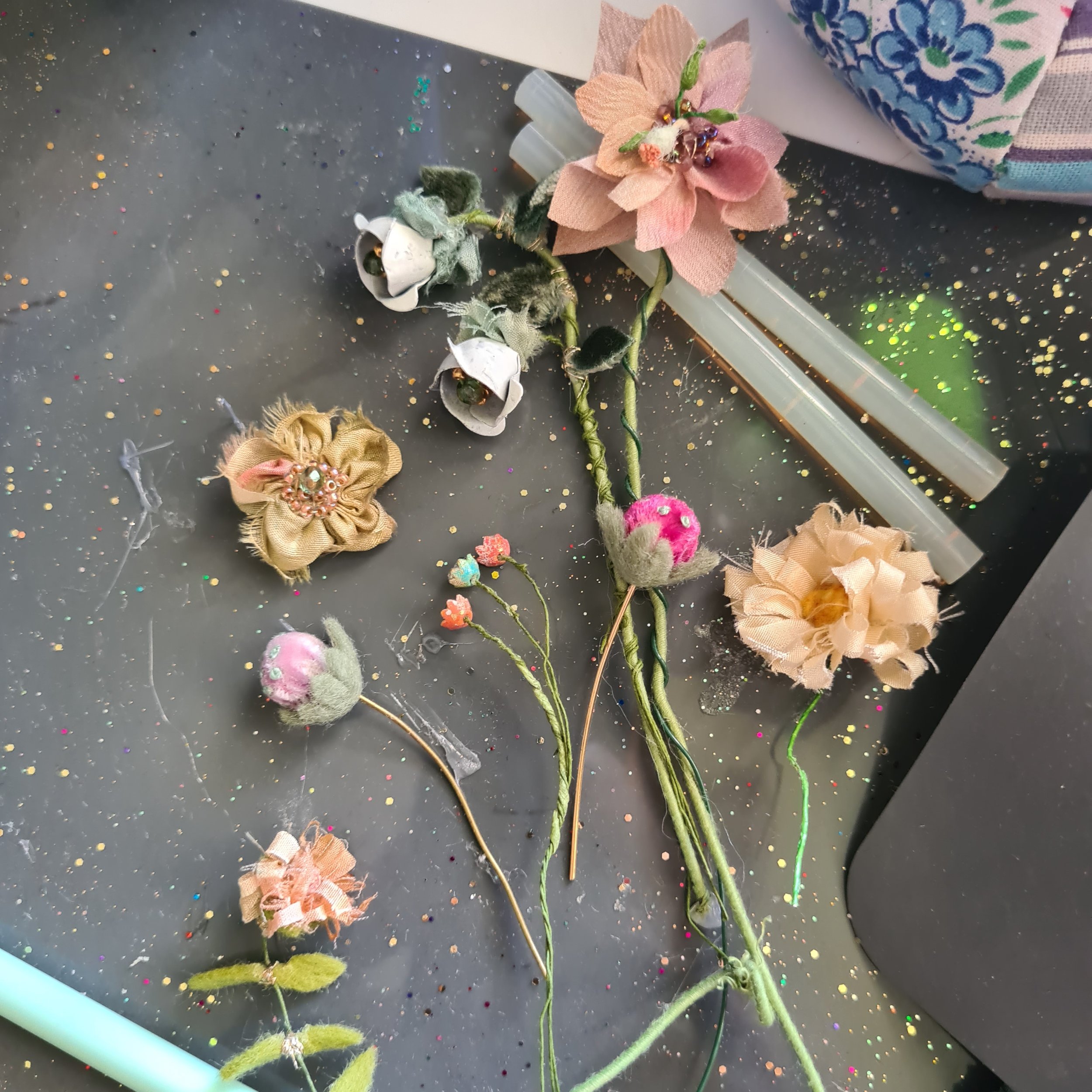 Craft materials for making fabric flowers, including pink, beige, and white fabric flowers with beads, felt, and wire stems, scattered on a black surface with decorative elements.