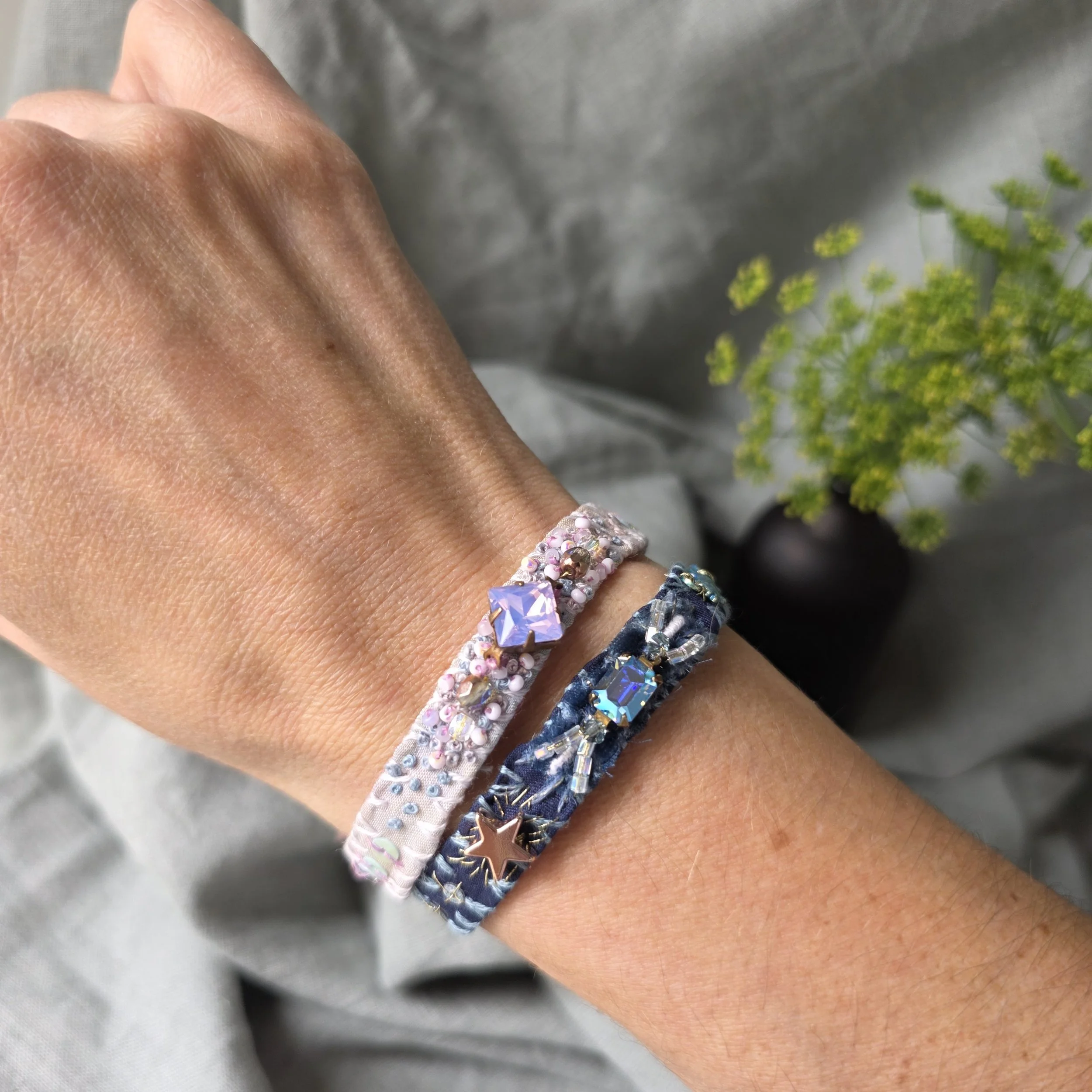 A person's wrist wearing two silk fabric bracelets with decorative beads and stones, one pink and one blue, with a small sparkling stone. In the background, there is a small black vase with green, tiny flowers.