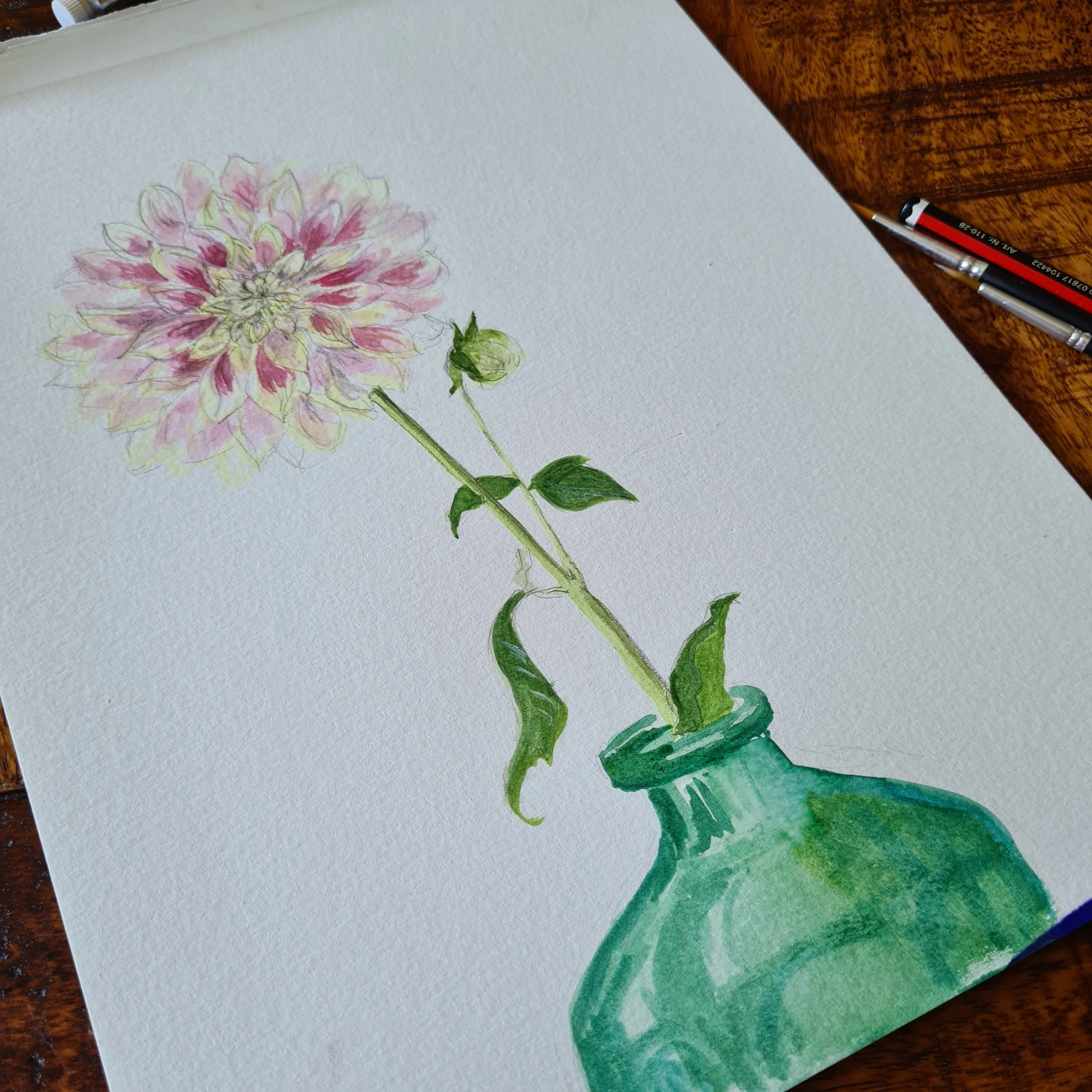 Watercolor painting of a pink and yellow dahlia flower in a green vase with a pencil on a wooden table.