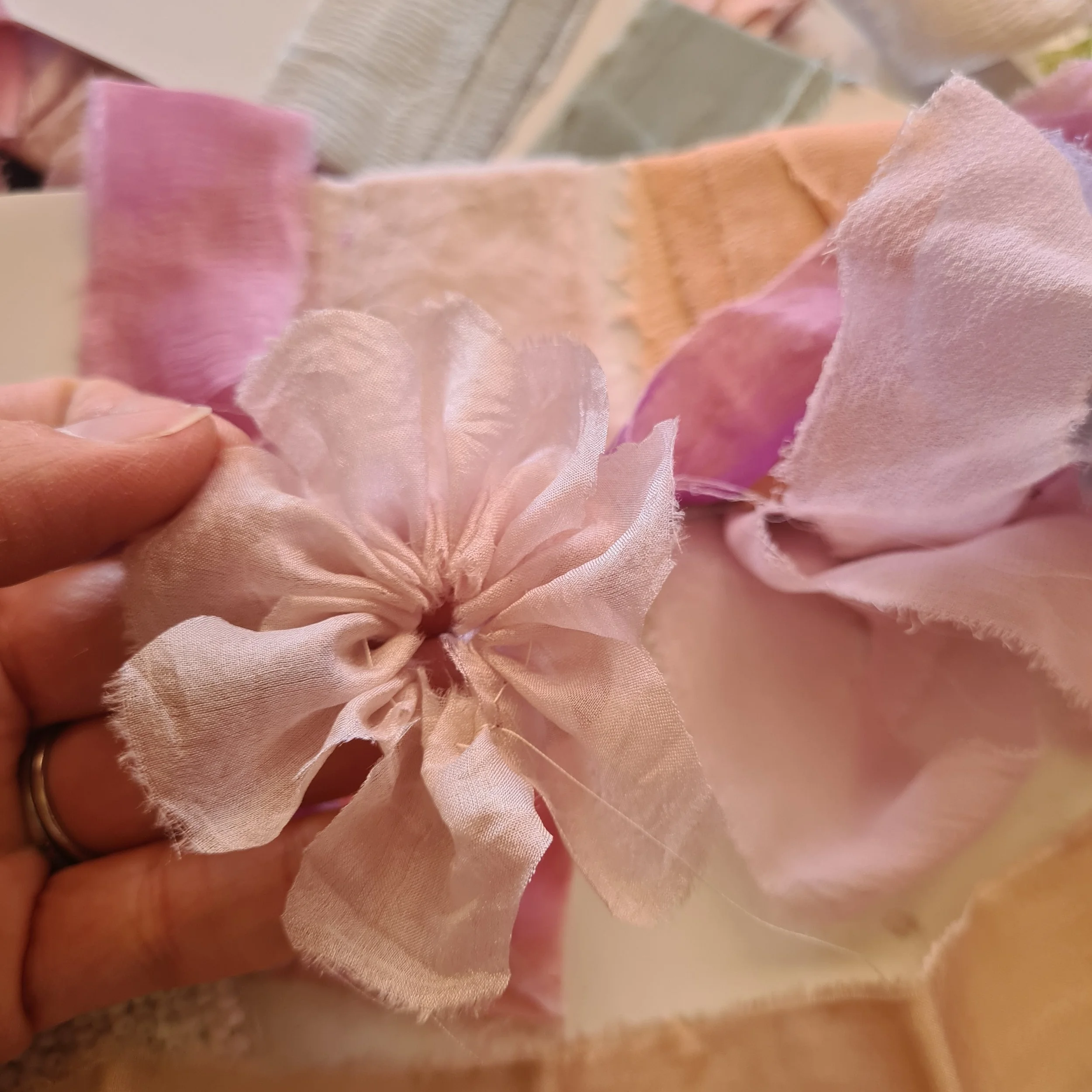 A hand holding a pink fabric flower with ruffled petals, set against a background of pink and beige fabric pieces.