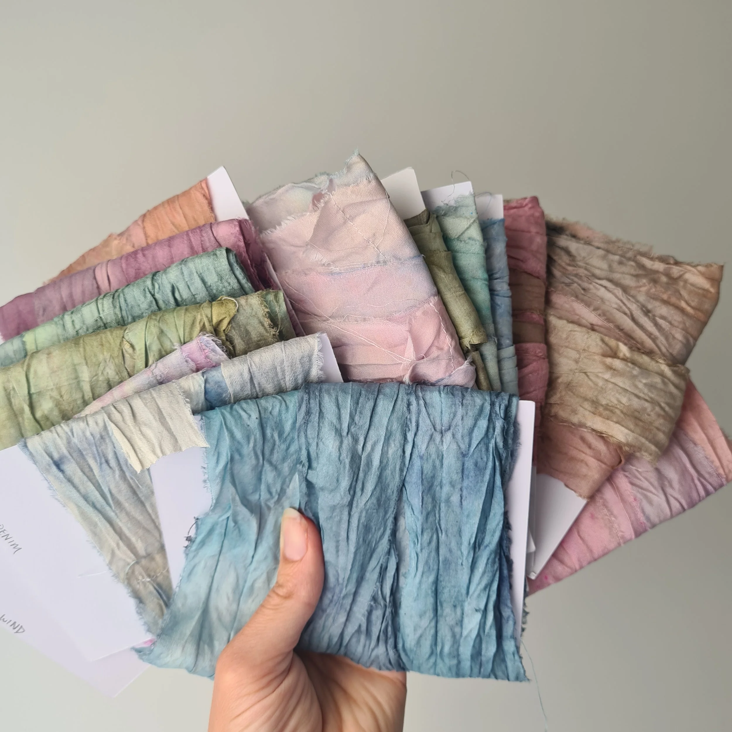 Hand holding a fan of silk ribbons on cards in various pastel colors with a textured, crinkled appearance.