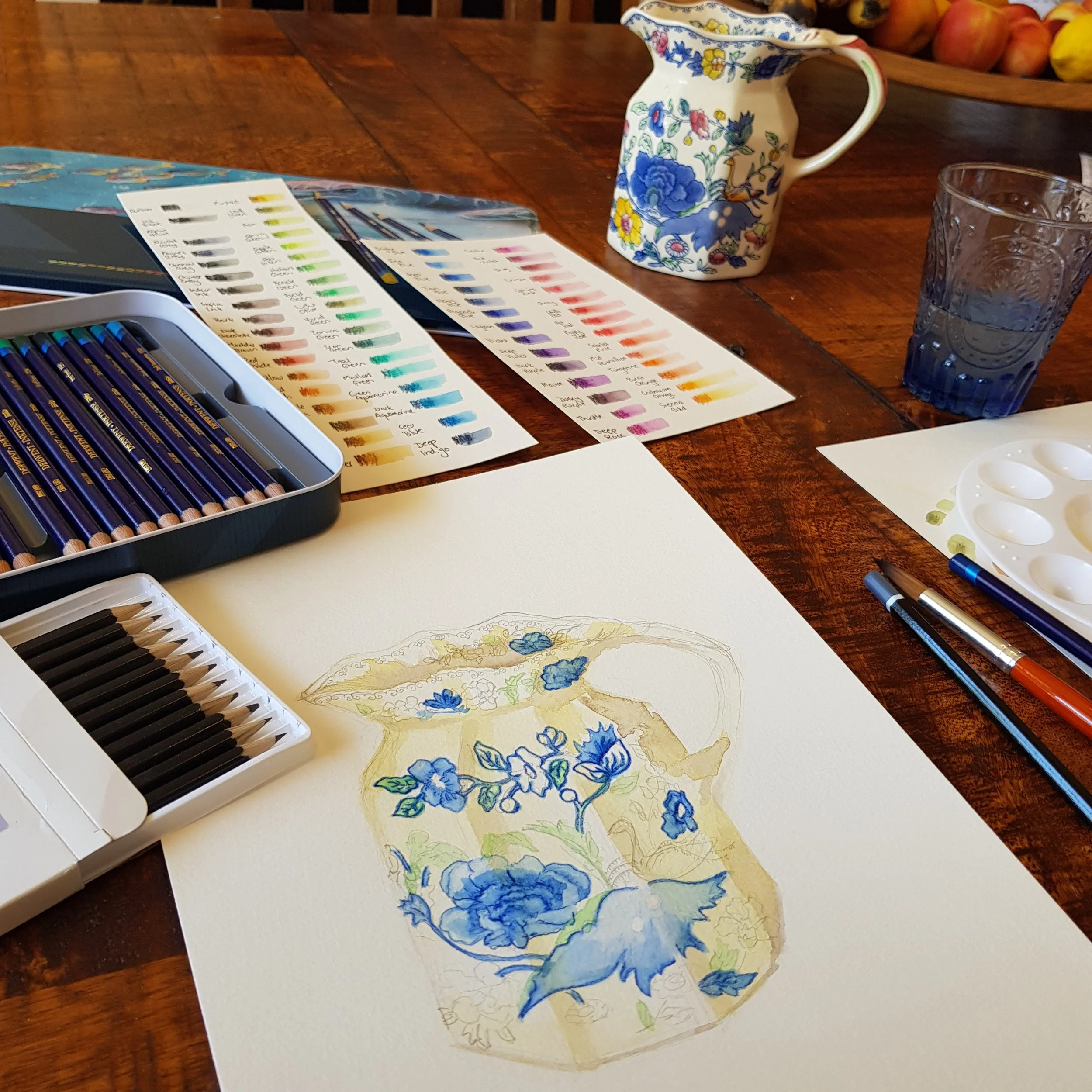 A watercolor painting of a blue floral vase on white paper, with watercolor paints, brushes, and color swatches on a wooden table.