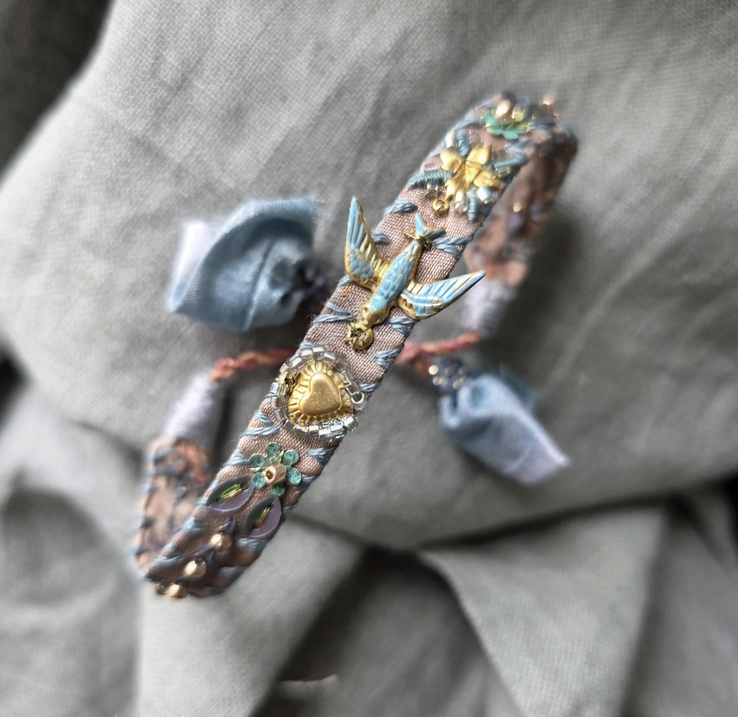 An embroidered silk bracelet in blues and greys with decorative embroidery including small bird, heart shape, and flower motifs, positioned on a grey textured fabric background.