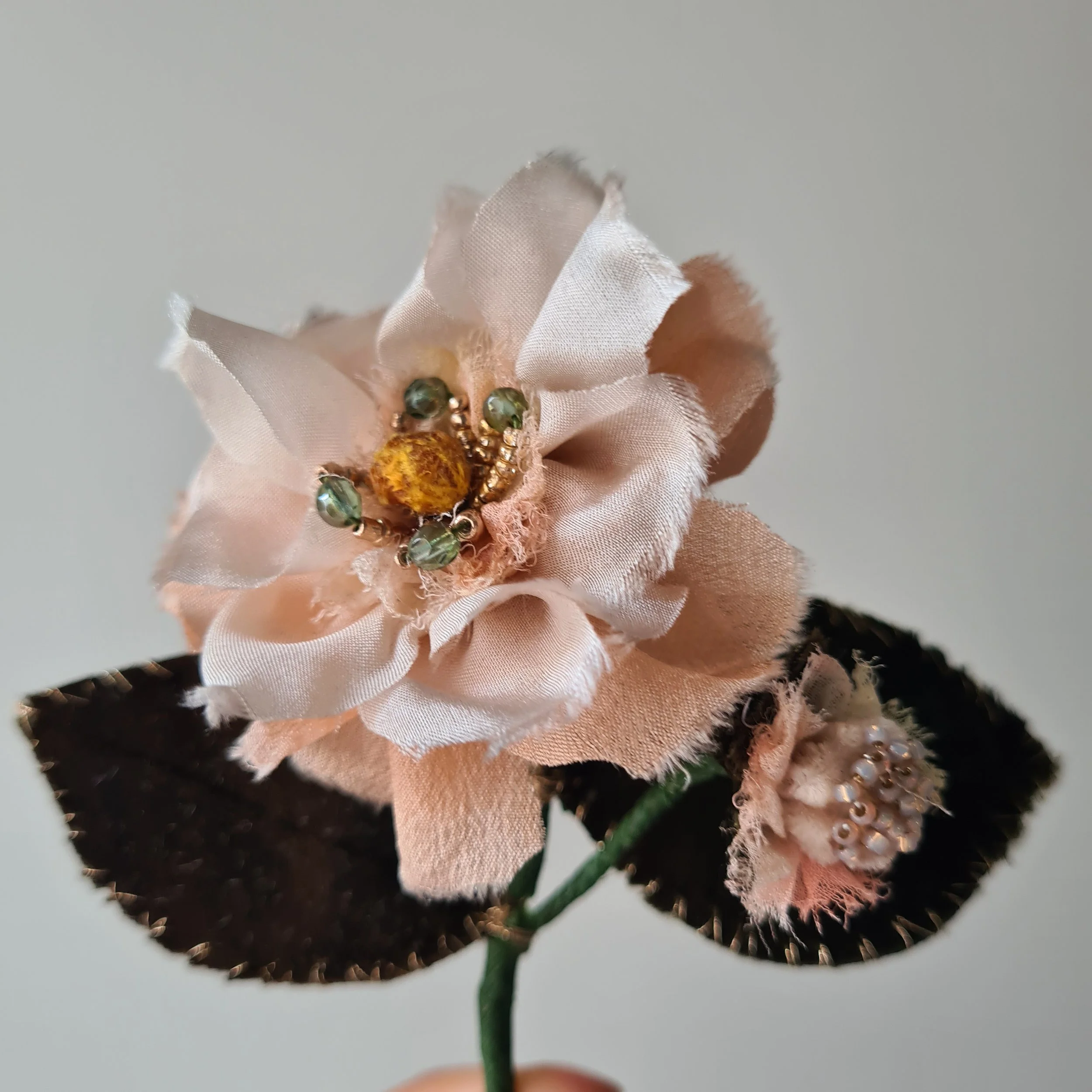 A silk and velvet fabric flower brooch with pink, peach and beige silk petals adorned with decorative beads, attached to dark green velvet leaves with a green stem.