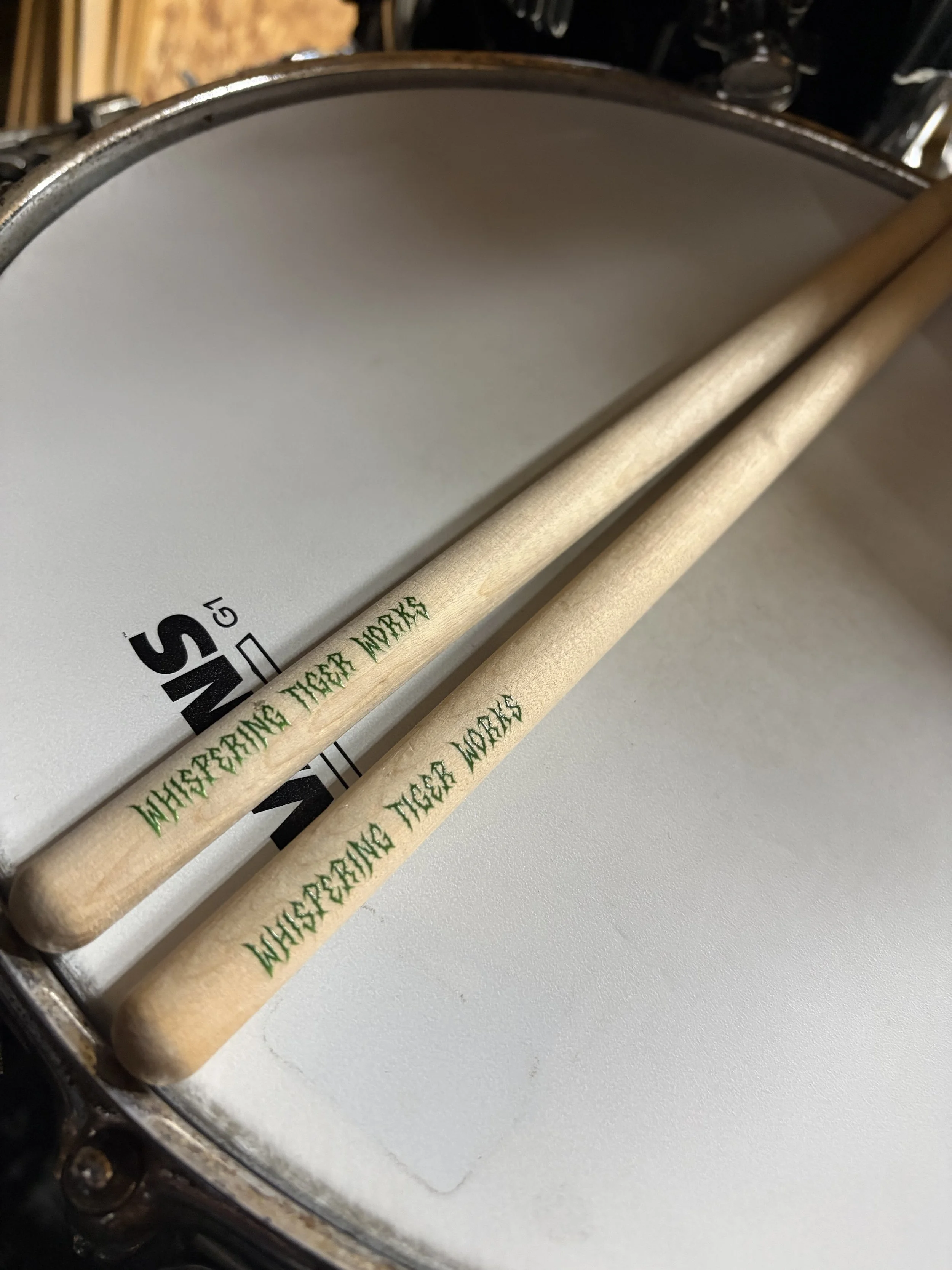 Close-up of a drum with a drumstick resting on the drumhead; the drumstick has green text that reads 'Whispering Tiger Works'.