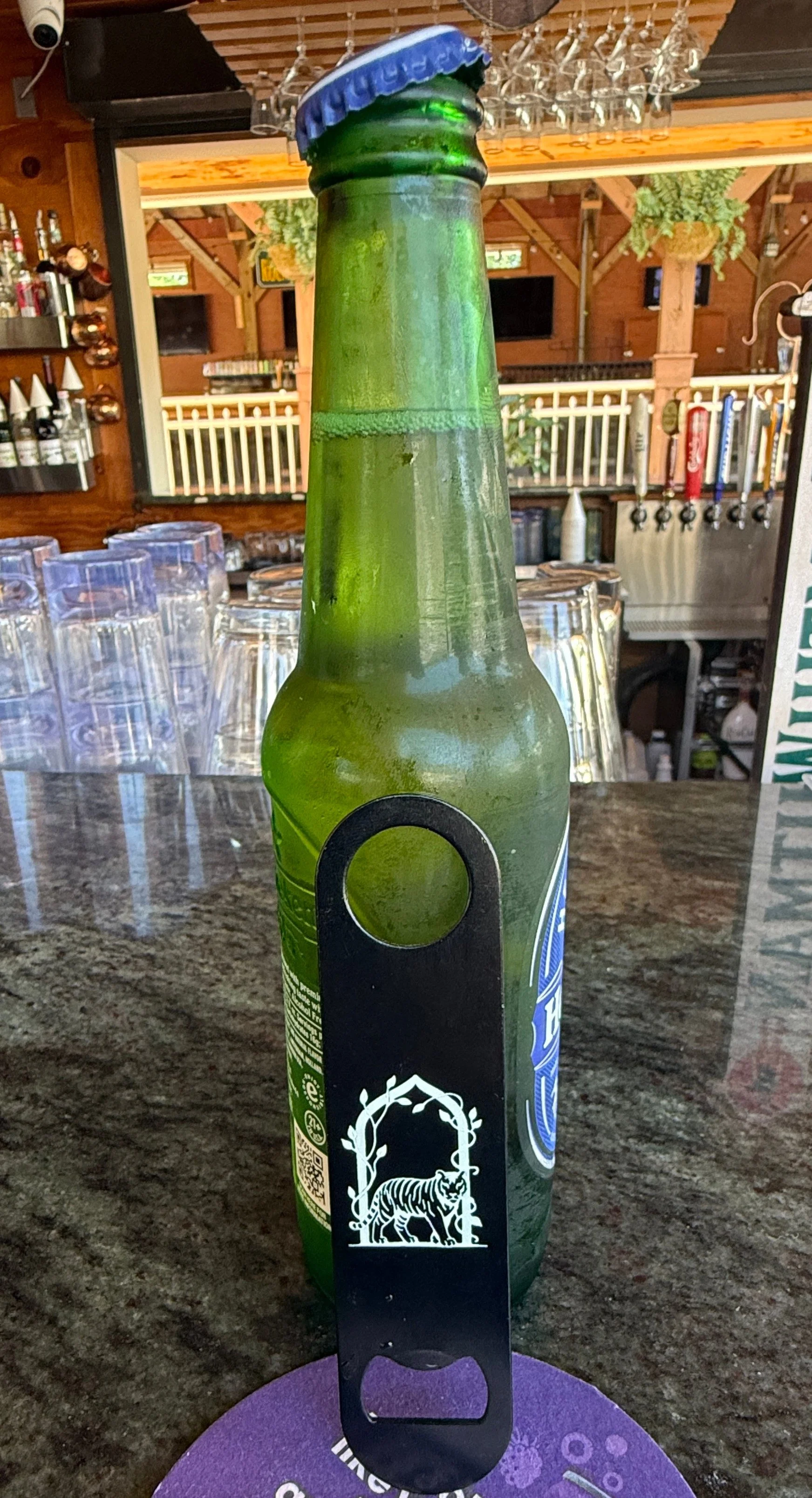 A green glass bottle with a black and white bottle opener featuring a tiger and an arch design, topped with a blue bottle cap, on a dark countertop in a bar or restaurant setting.