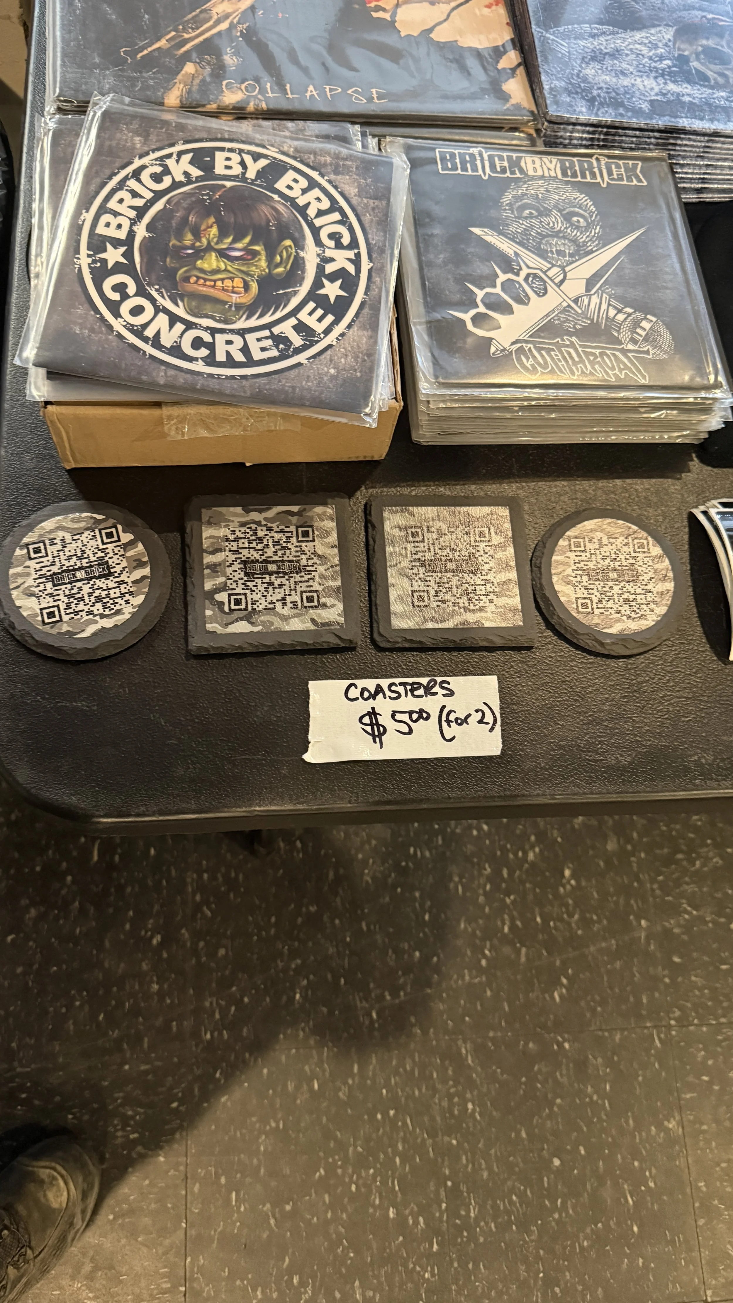 Stack of coasters with QR codes and illustrations, displayed on a table with a handwritten sign indicating price of $5 for two.