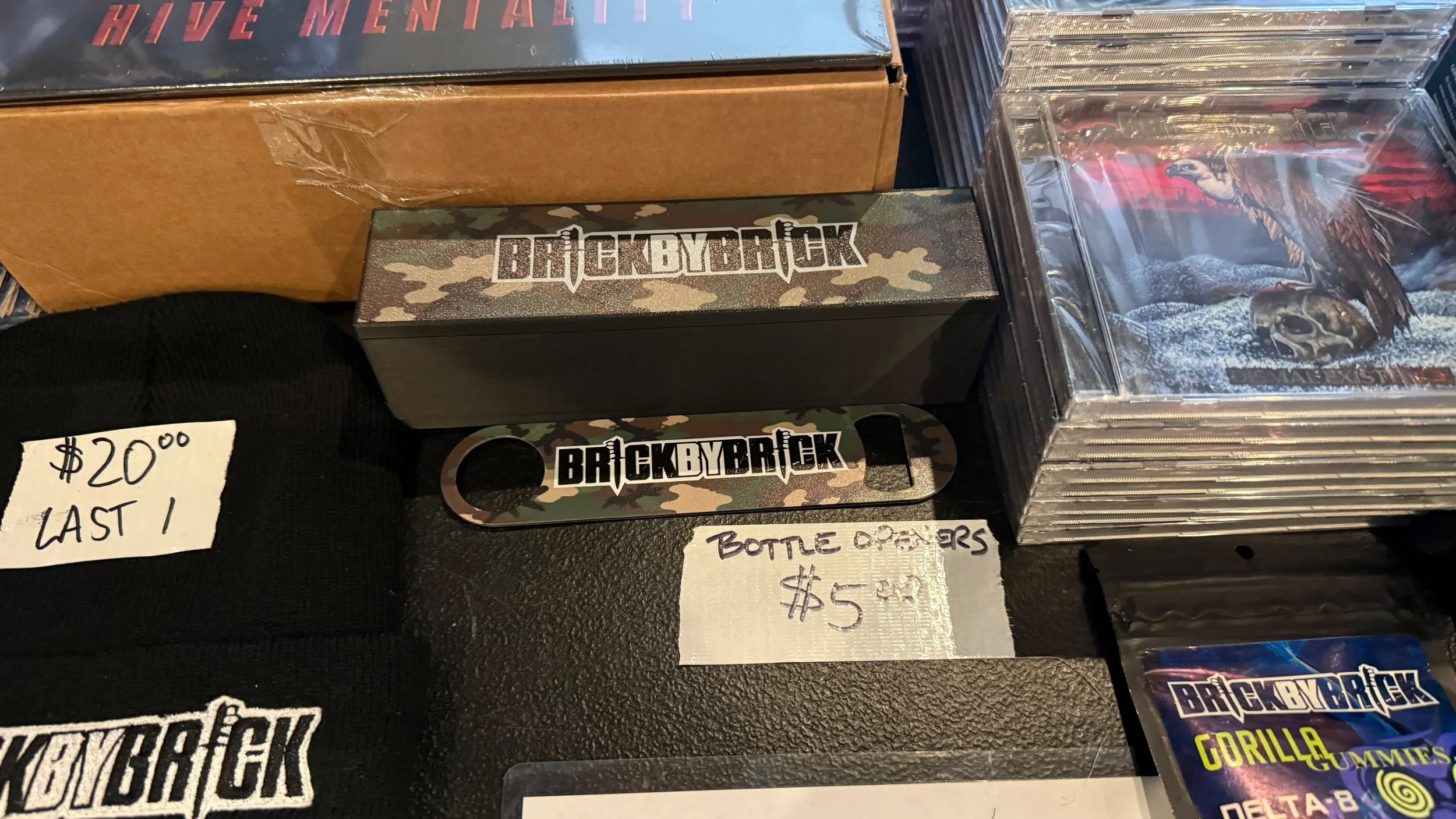 Various merchandise including a box labeled 'HIVE MENTAL' and a sticker with 'BRICK BY BRICK' in camouflage design, placed on a table with handwritten price tags and CD cases, some featuring fantasy or sci-fi artwork.