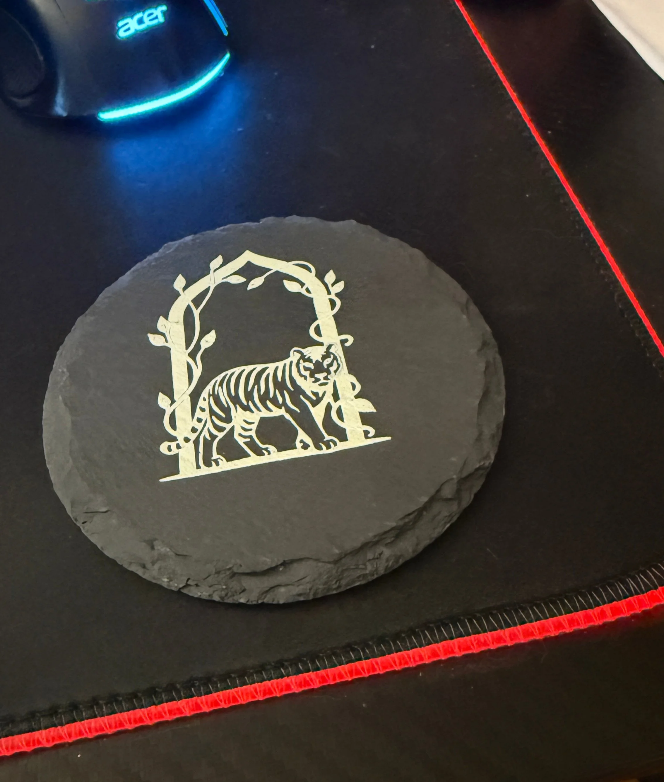 A round black stone with a white tiger illustration surrounded by branches, placed on a black surface with red stitching, near a black Acer remote control with a blue-lit power button.