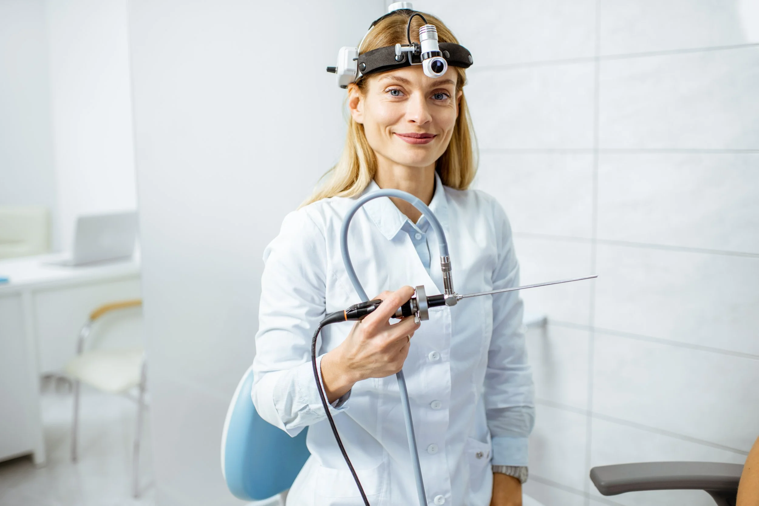 A female doctor wearing a white coat and a headlamp with a magnifier, holding and preparing to use a medical endoscope in a clinical setting.
