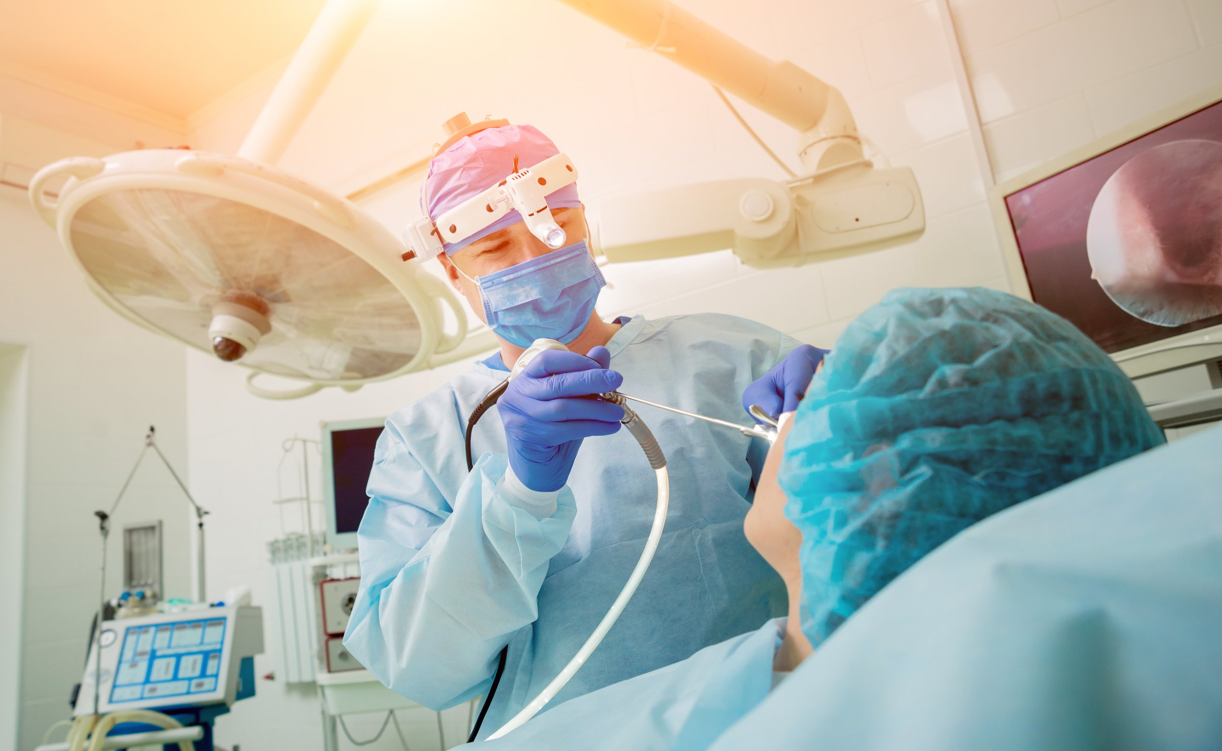 Surgeon performing endoscopic procedure in an operating room, wearing surgical mask, gown, gloves, and headlight, with medical equipment and monitors in the background.
