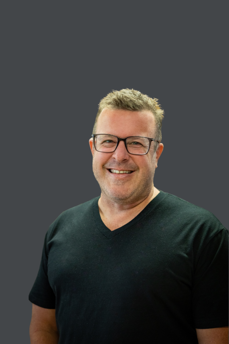 A smiling man with glasses, wearing a black V-neck shirt, standing in front of brown wooden blinds.