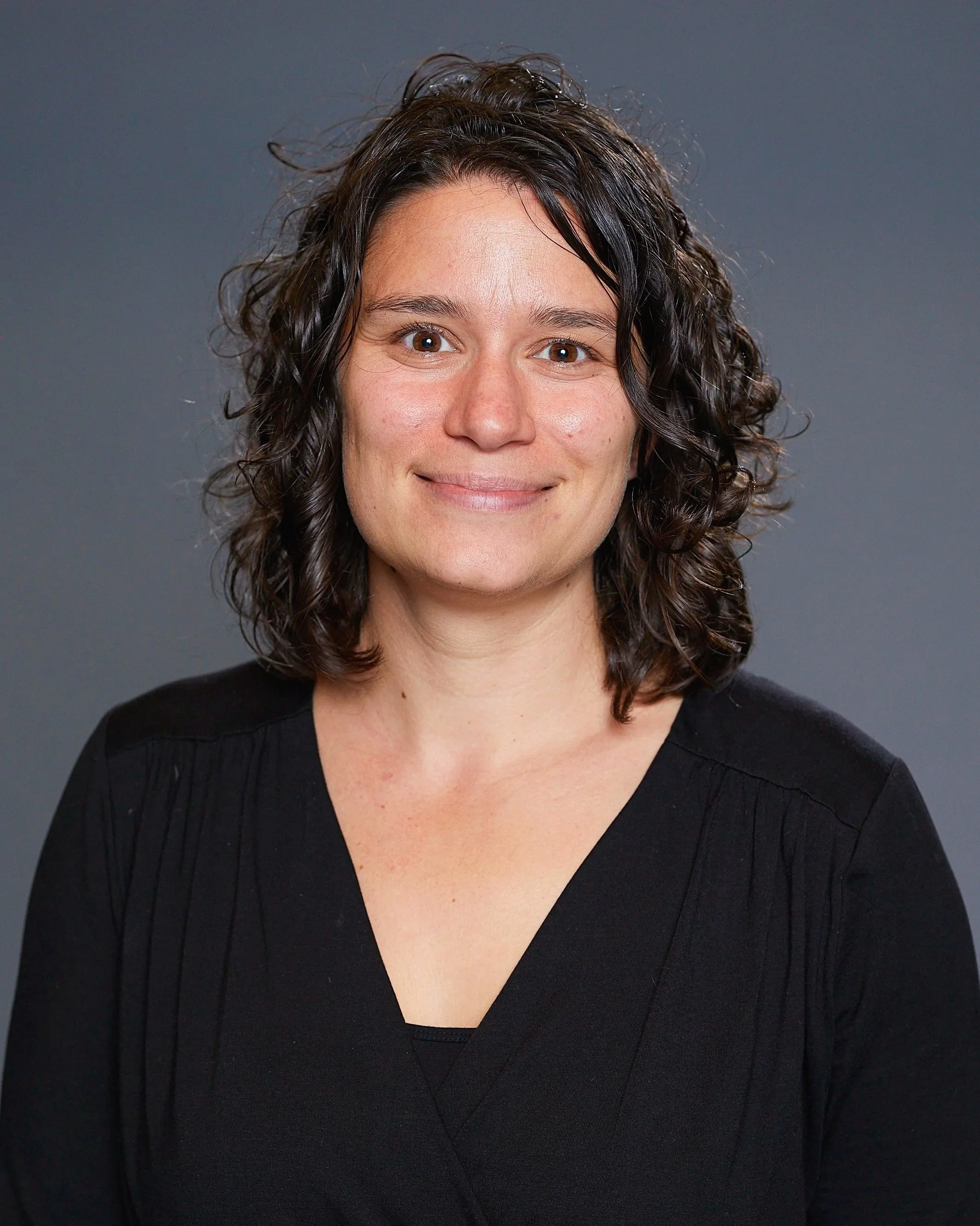 Close-up of a woman with dark curly hair wearing a black V-neck sweater, smiling, against a plain gray background.