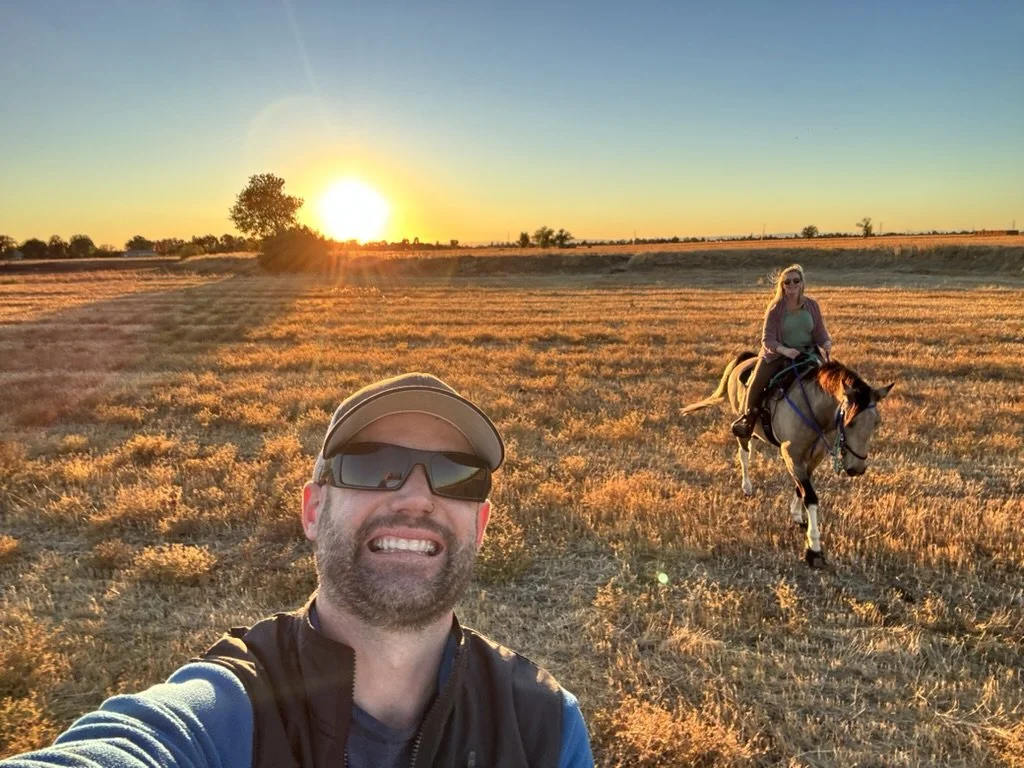 A man taking a selfie in a wide open field during sunset with a woman riding a horse in the background.