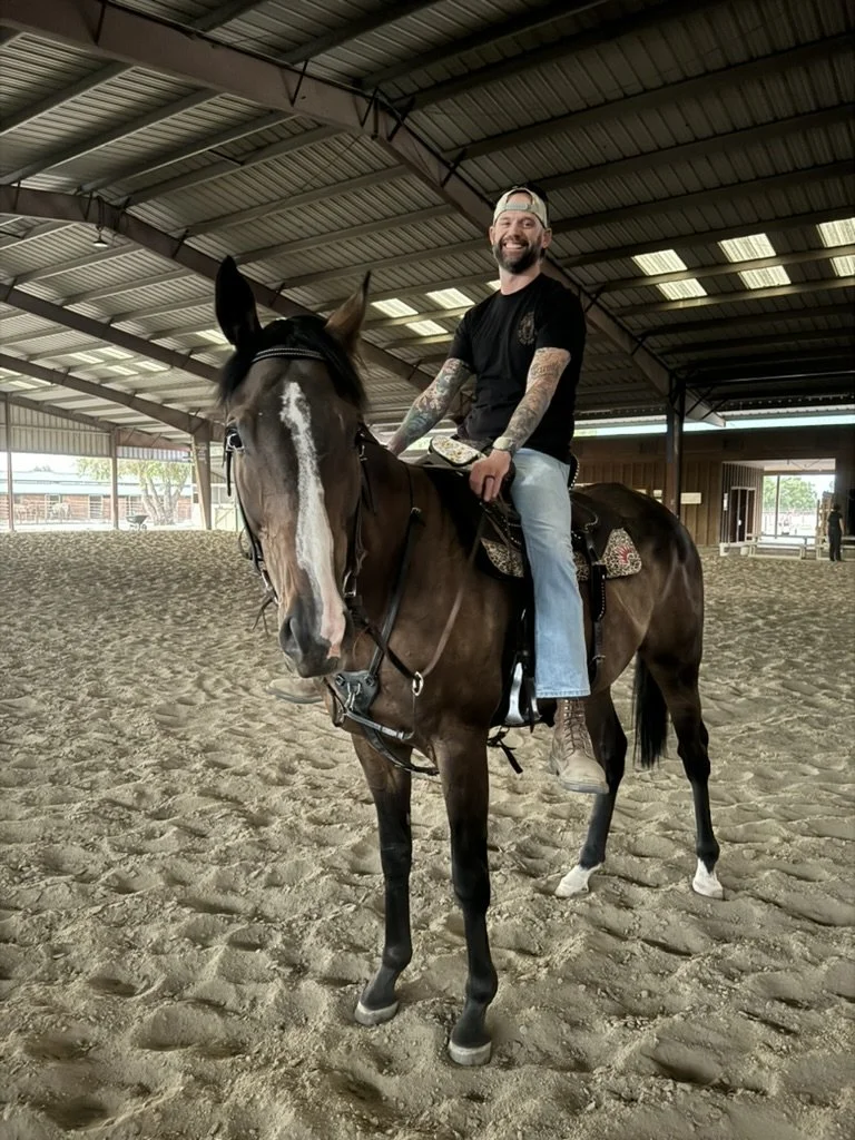 A man with a beard, tattoos, and wearing a black T-shirt, blue jeans, and a backwards baseball cap is riding a brown horse inside an indoor riding arena. The man is smiling and sitting on the horse's saddle.