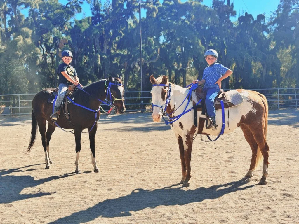 Two young boys wearing helmets riding horses in an outdoor riding arena with trees in the background.