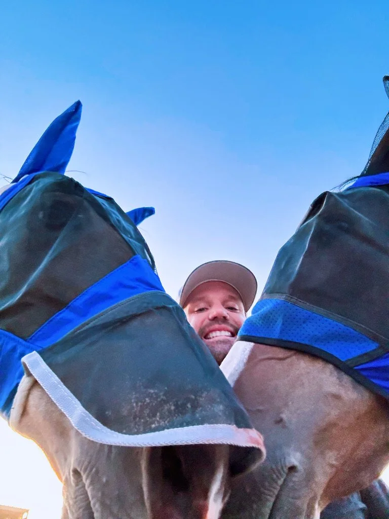 A person smiling at the camera, seen from below, with two horses on either side and a blue sky above.