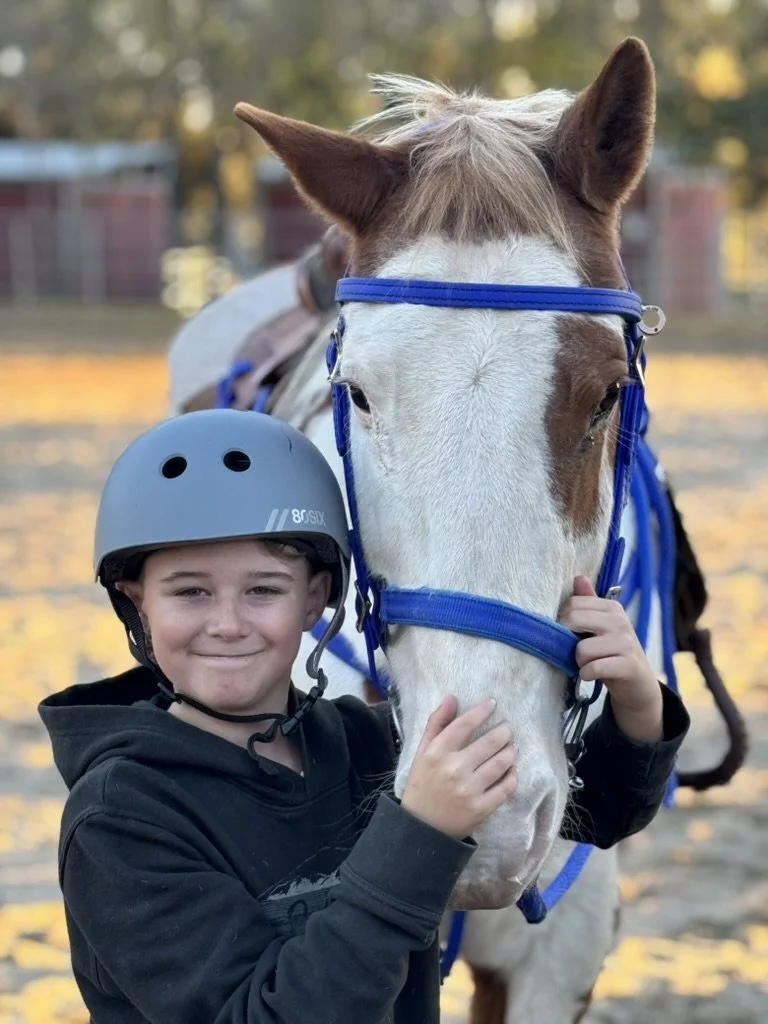 A smiling boy in a black hoodie wearing a riding helmet, petting a white horse with brown patches and a blue halter.