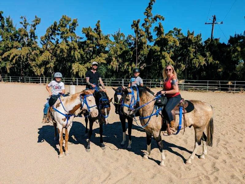 Four people riding horses in an outdoor riding arena with a sandy surface, surrounded by a fence and lush green trees under a clear blue sky.