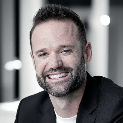 A smiling man with short dark hair and a beard, wearing a black blazer and white shirt, in a professional setting.