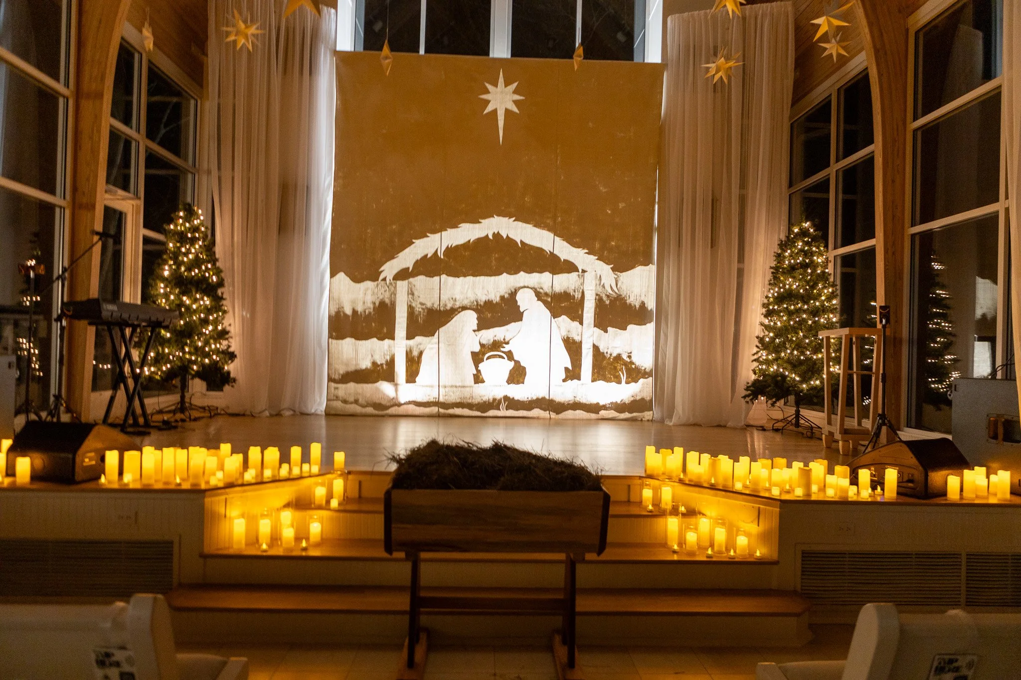 Nativity scene tableau with silhouettes of Mary, Joseph, and baby Jesus under a star, backdrop with Christmas trees, candles along the front, and star decorations hanging from the ceiling.