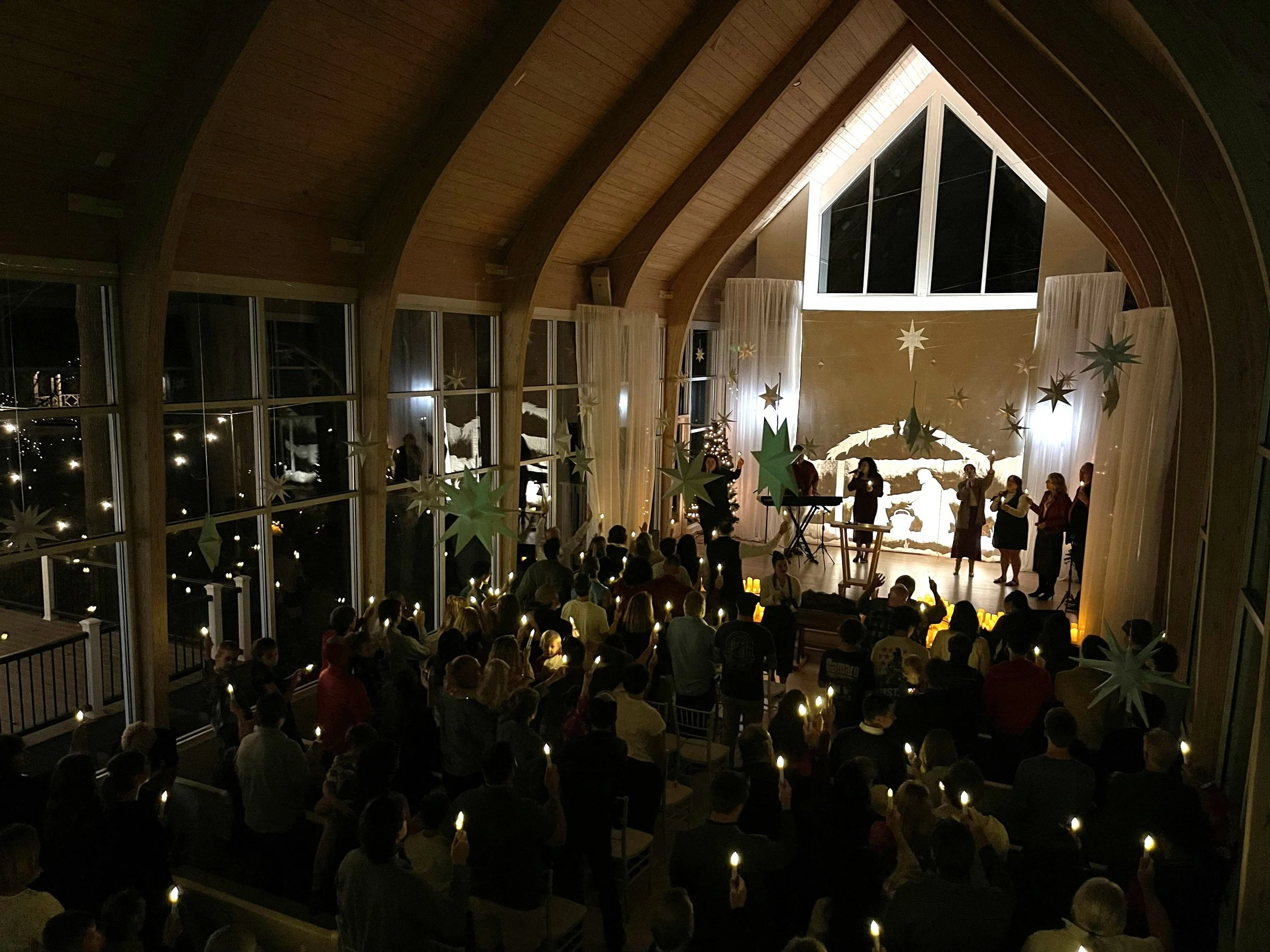 A Christmas service inside a church with many people holding candles, a Christmas tree, a nativity scene, and choir singers performing on stage.