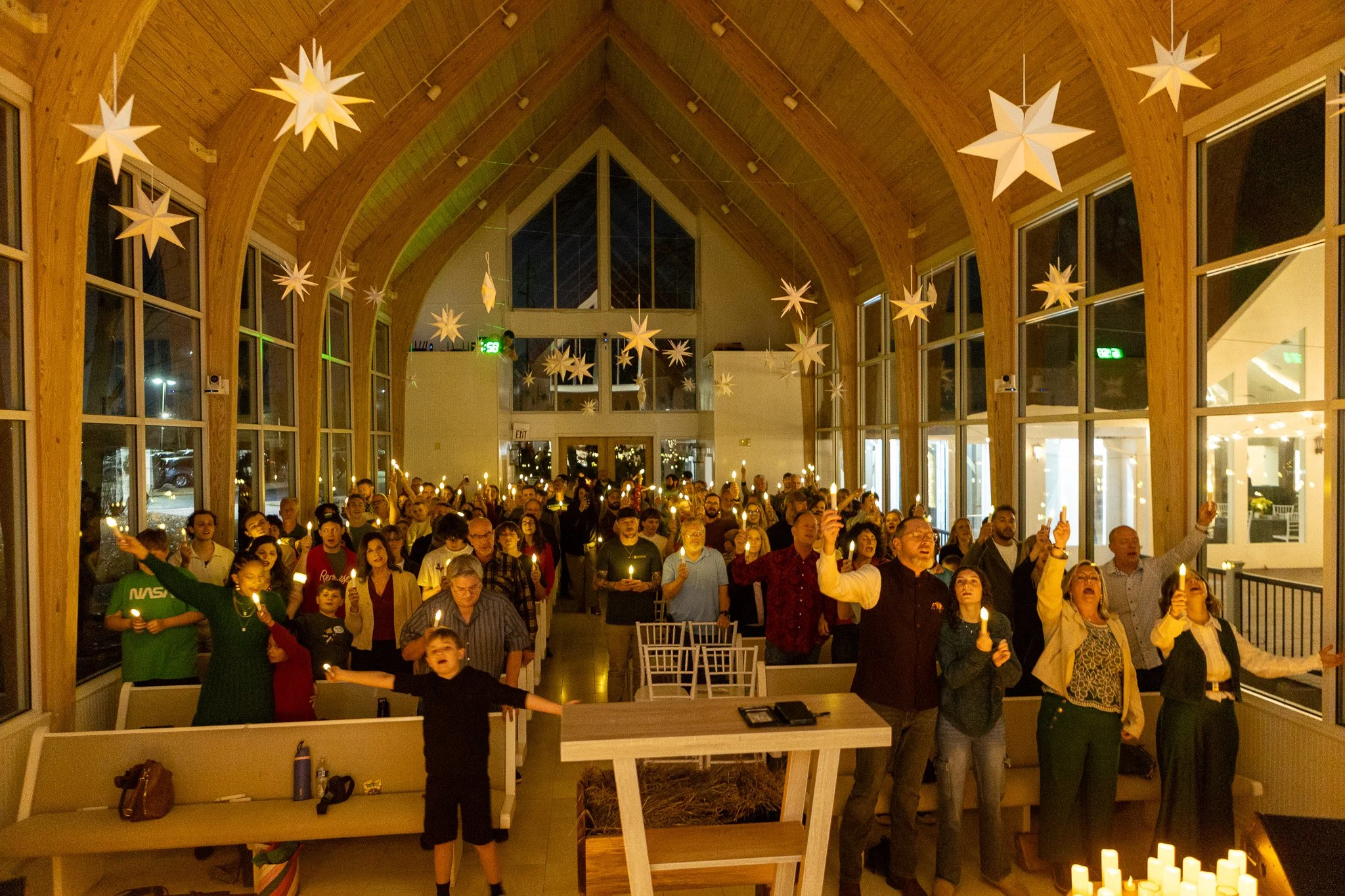 People gathered inside a church for a candlelight service. The church has wooden arches and star-shaped decorations hanging from the ceiling. Attendees are holding candles, standing reverently, with some raising their hands in worship.