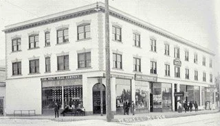 Black and white photo of a three-story building on a city street corner, with large windows and storefronts on the ground level.