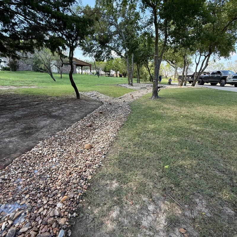 A gravel path flowing through a grassy park area with trees, with parked trucks and people in the background.