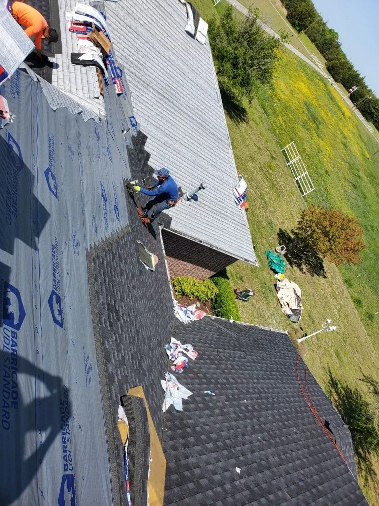 Roof construction workers installing shingles on a sloped roof, with roofing materials, tools, and a partially completed section visible.