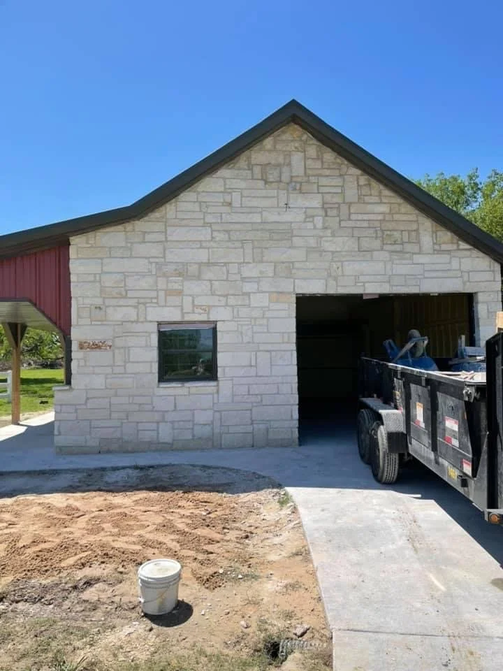 Newly constructed garage with an open door, a black trailer parked outside, and a small window on the left wall. The building has a stone exterior with a dark-colored roof.