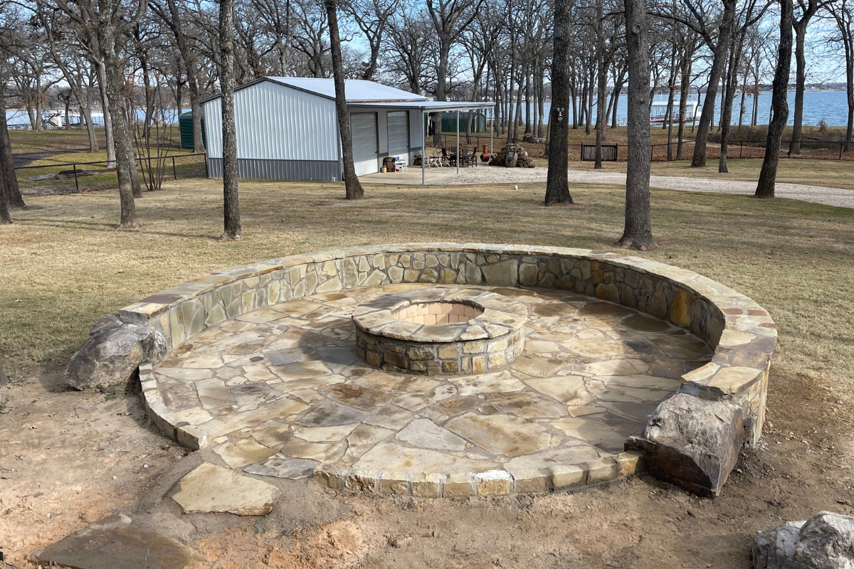 A circular stone fire pit with a surrounding stone bench in a park with trees, a lake, and a building in the background.