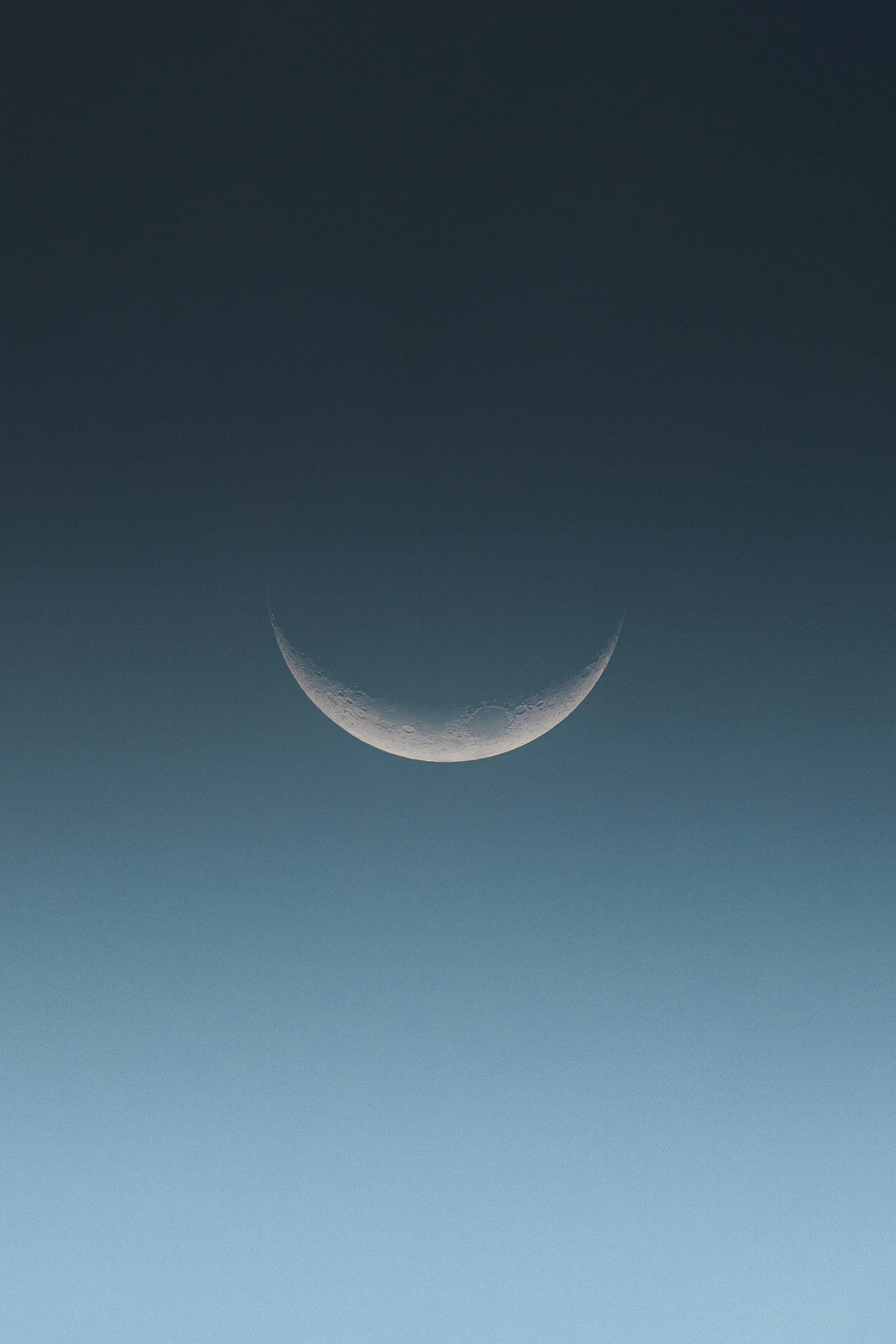 Close-up photograph of a waxing crescent moon in the early evening sky, showing detailed craters.