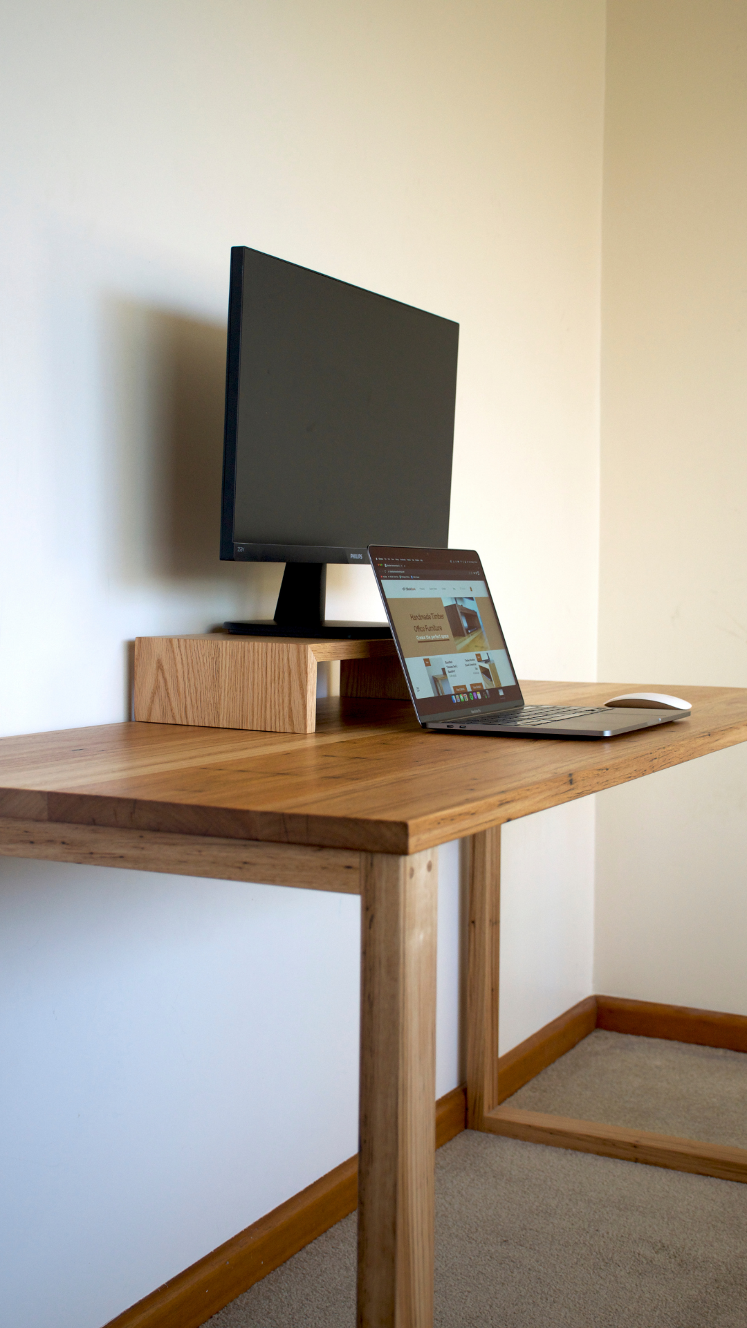 A desktop setup with a computer monitor, a laptop displaying a website, and a wireless mouse on a wooden desk against a beige wall.
