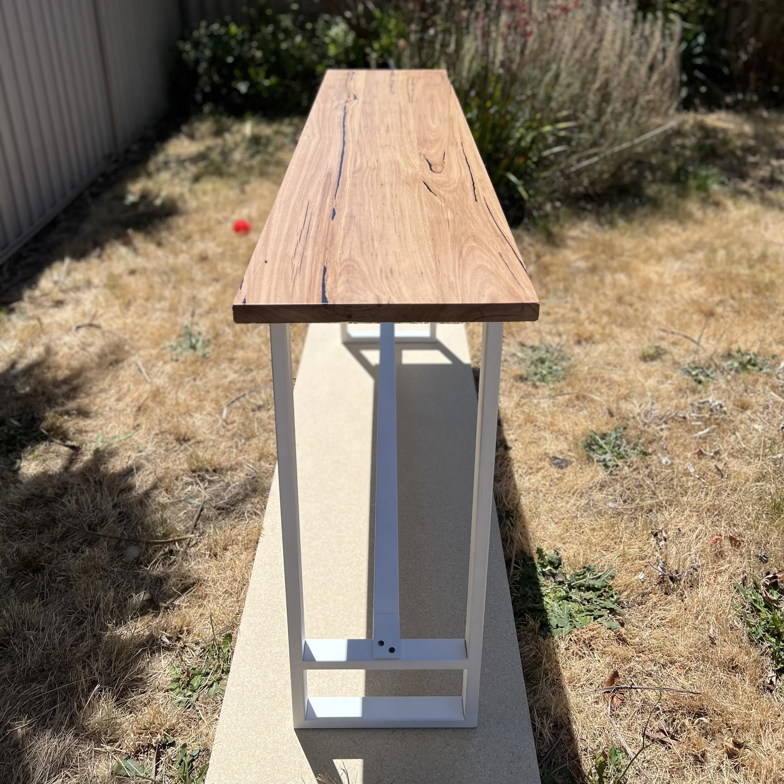 A long wooden table with a natural finish and visible grain, supported by a white metal frame, situated outdoors on a concrete pathway in a yard with brown grass and some greenery.