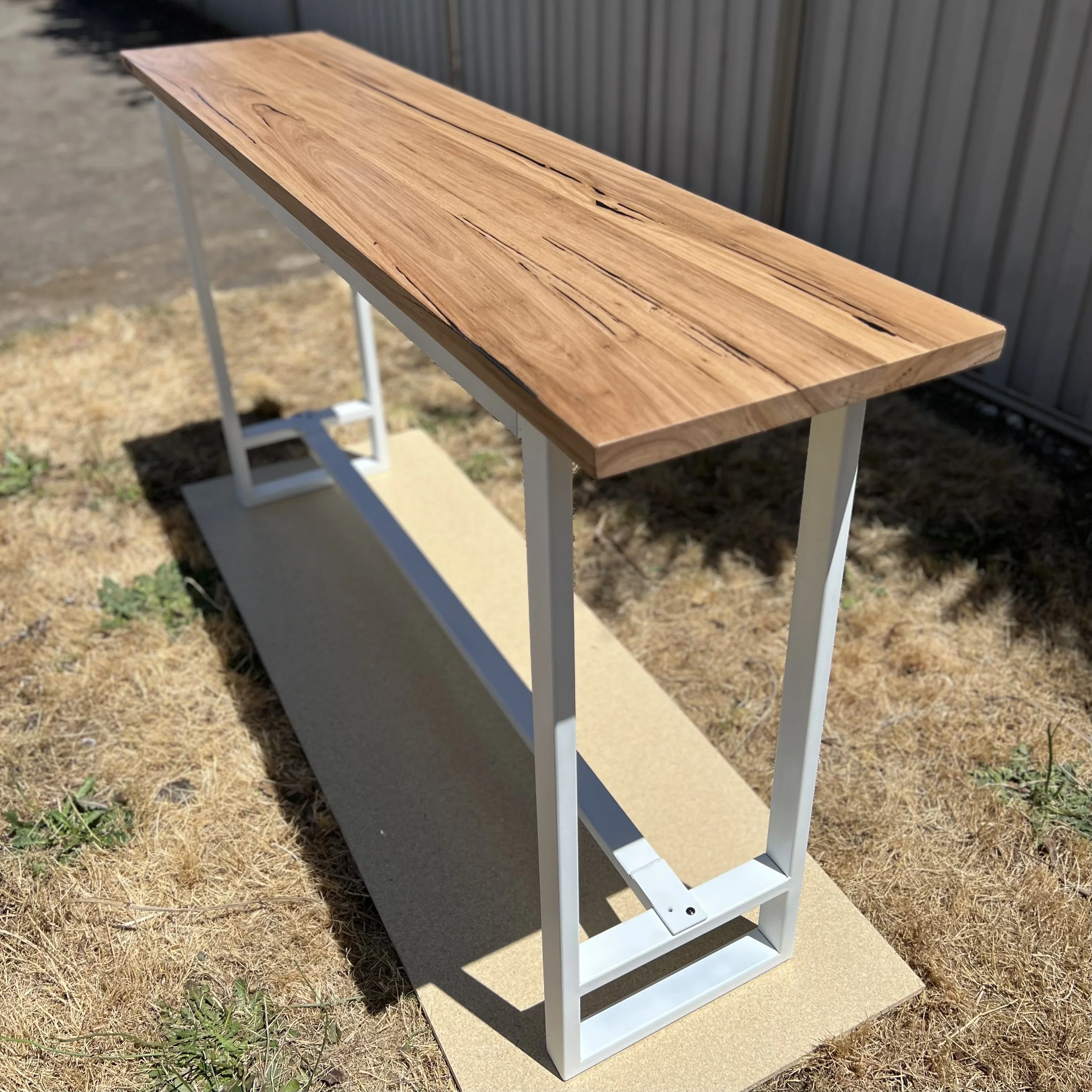 Wooden top table with white metal frame outdoors on grassy ground.